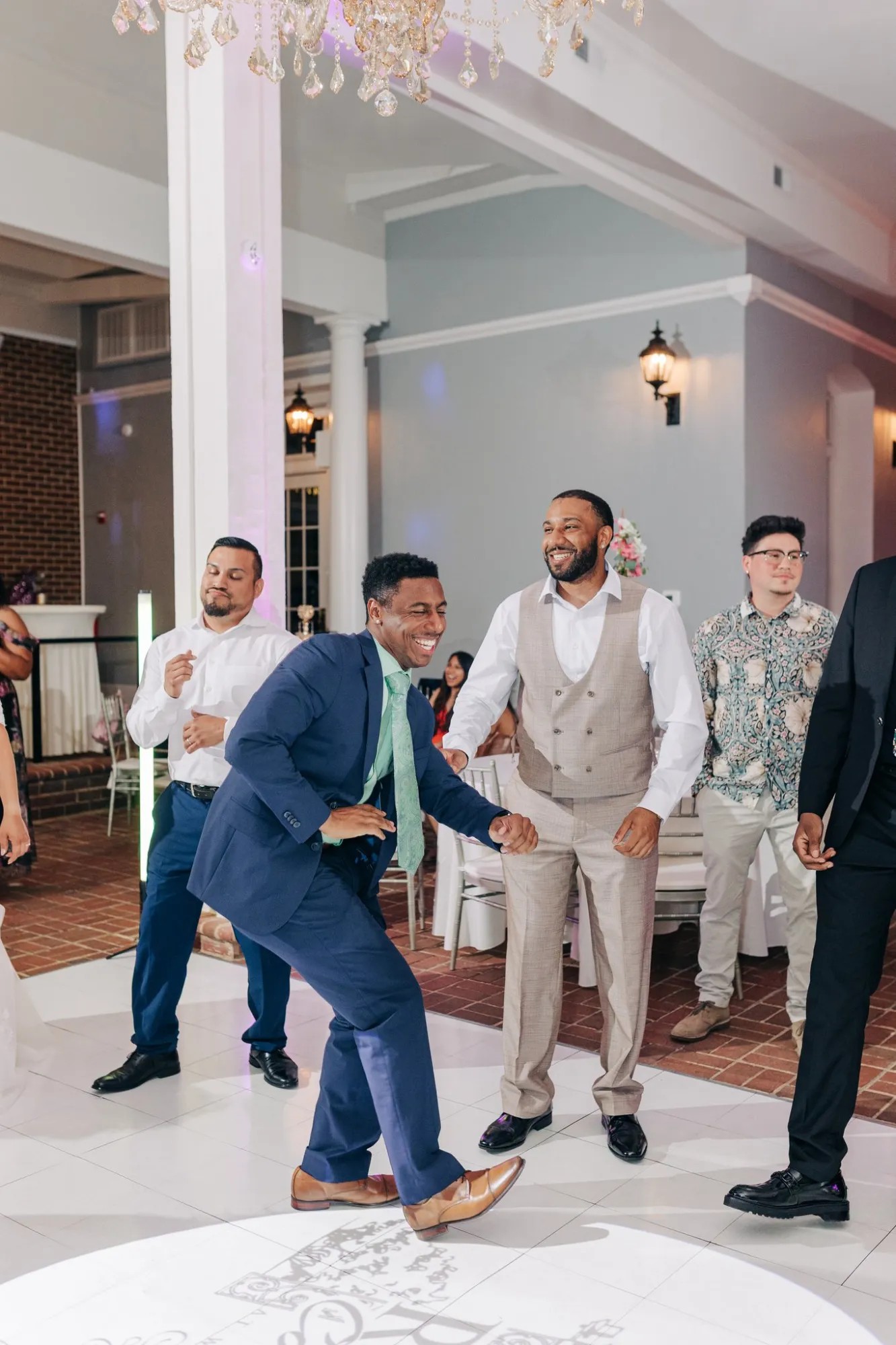 A group of men dressed in formal and semi-formal attire dancing and smiling indoors on a white tiled floor with a chandelier overhead and soft wall lighting.