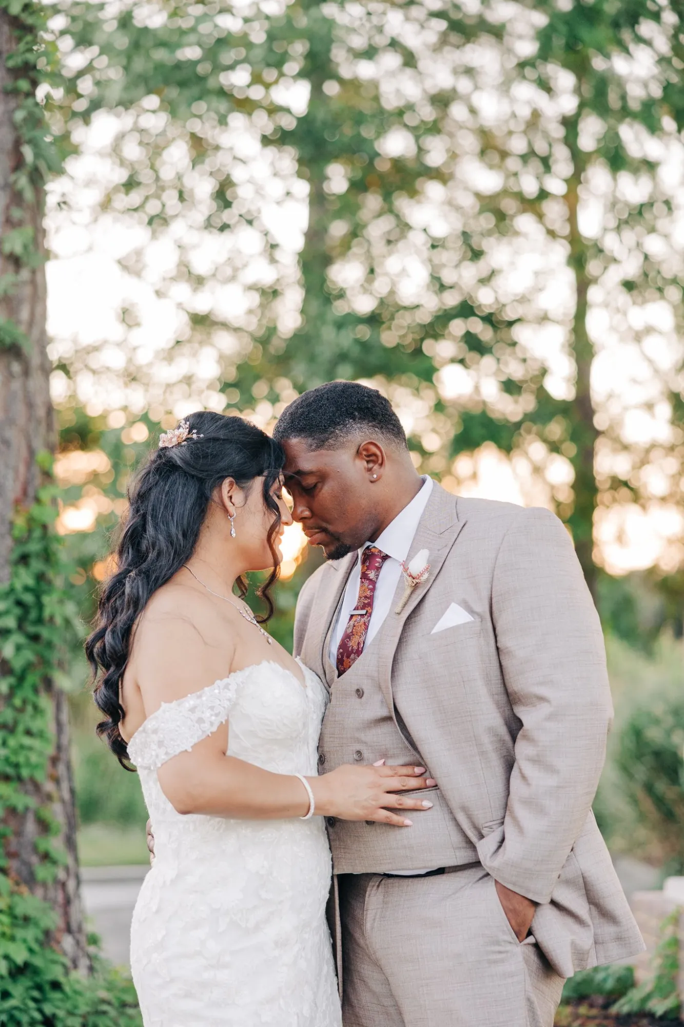 Bride in a white lace off-shoulder wedding dress and groom in a beige suit with floral tie stand close with their foreheads touching outdoors near trees at sunset.