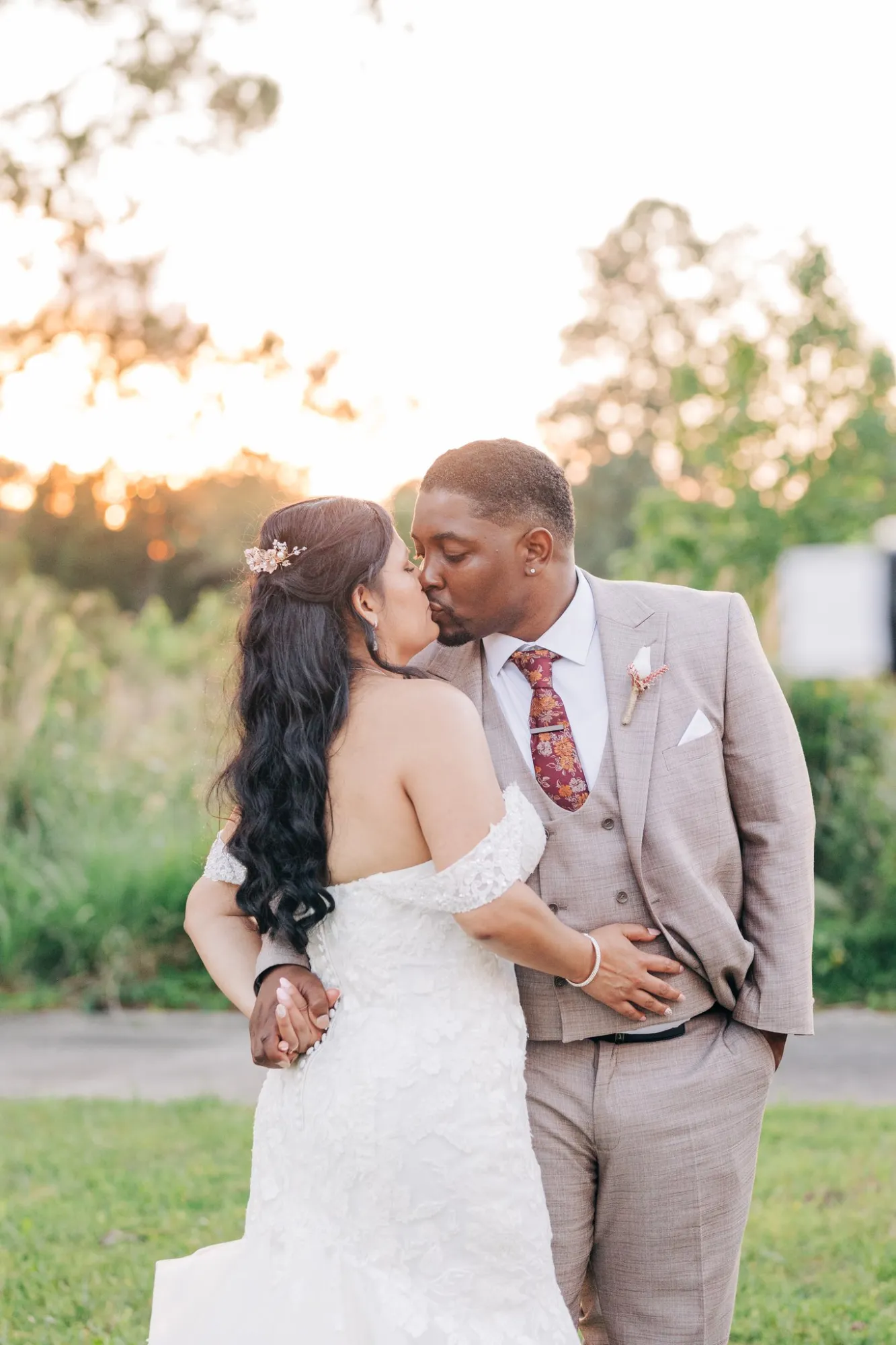 A bride and groom kiss outdoors at sunset; the bride wears an off-shoulder white lace wedding dress, and the groom wears a light brown suit with a floral tie.