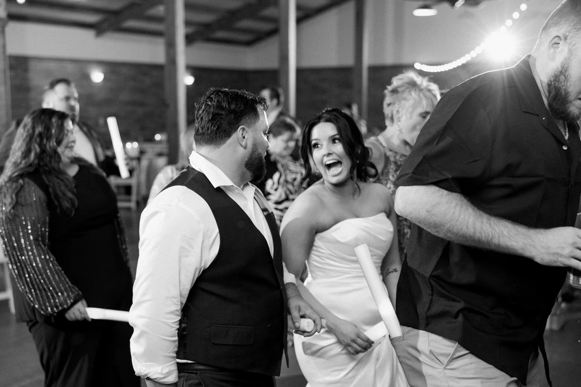 A black and white photo of a bride in a strapless wedding dress laughing while talking to a man in a vest at a lively indoor wedding reception with other guests in the background.