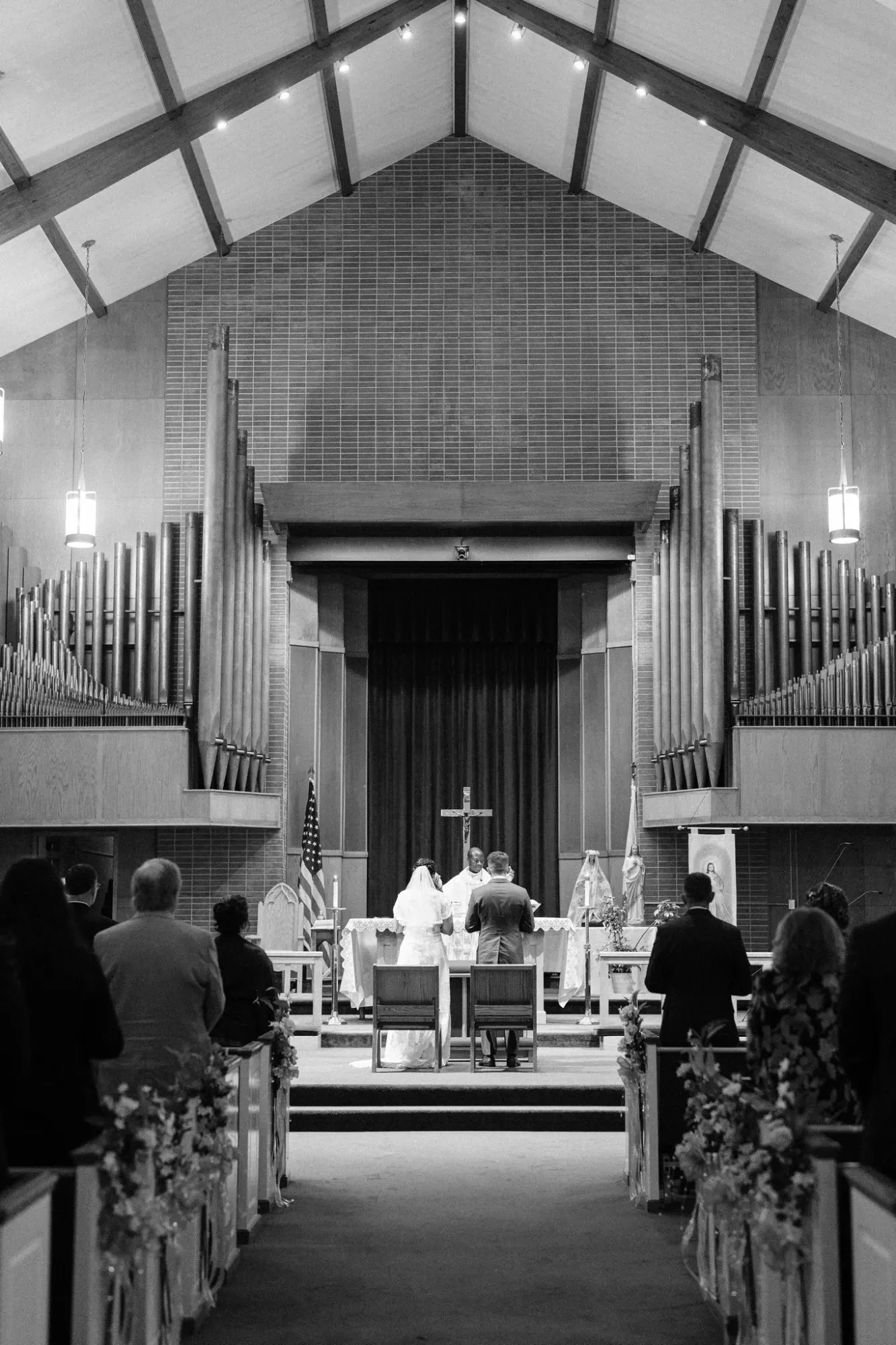 Black and white photo of a wedding ceremony inside a church, showing the bride and groom standing at the altar with a priest and guests seated in pews decorated with flowers.