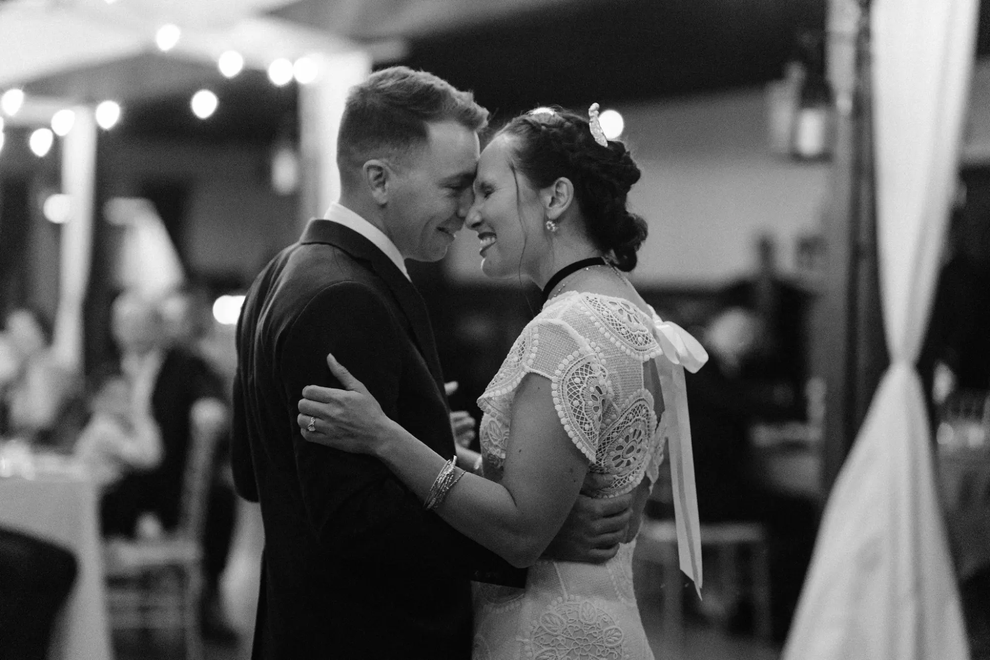 Black and white photo of a bride and groom dancing closely and smiling at each other at their wedding reception, with blurred guests and string lights in the background.