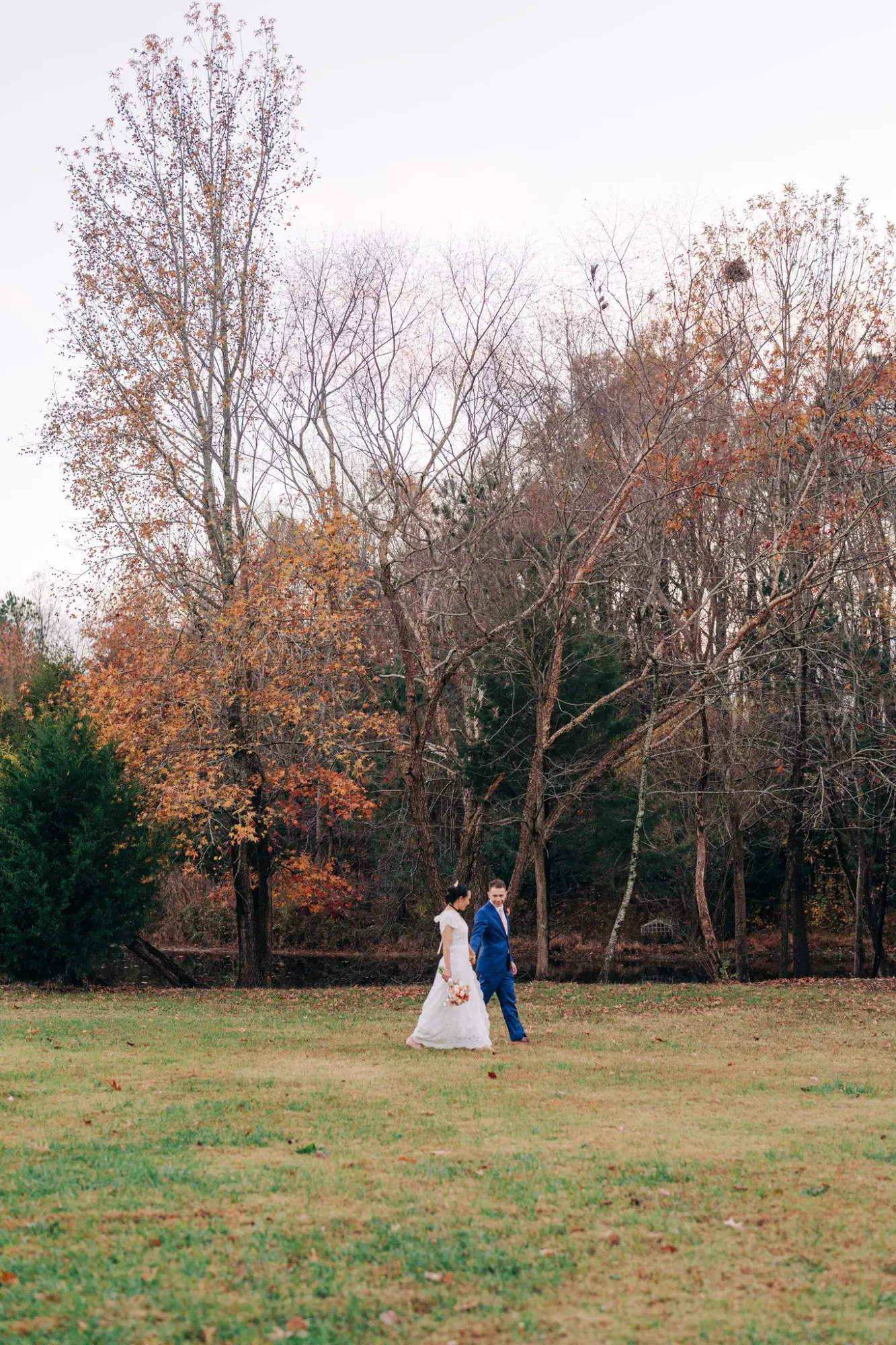 A bride in a white wedding dress holding a bouquet and a groom in a blue suit walk side by side on a grassy field with bare and autumn-colored trees in the background.