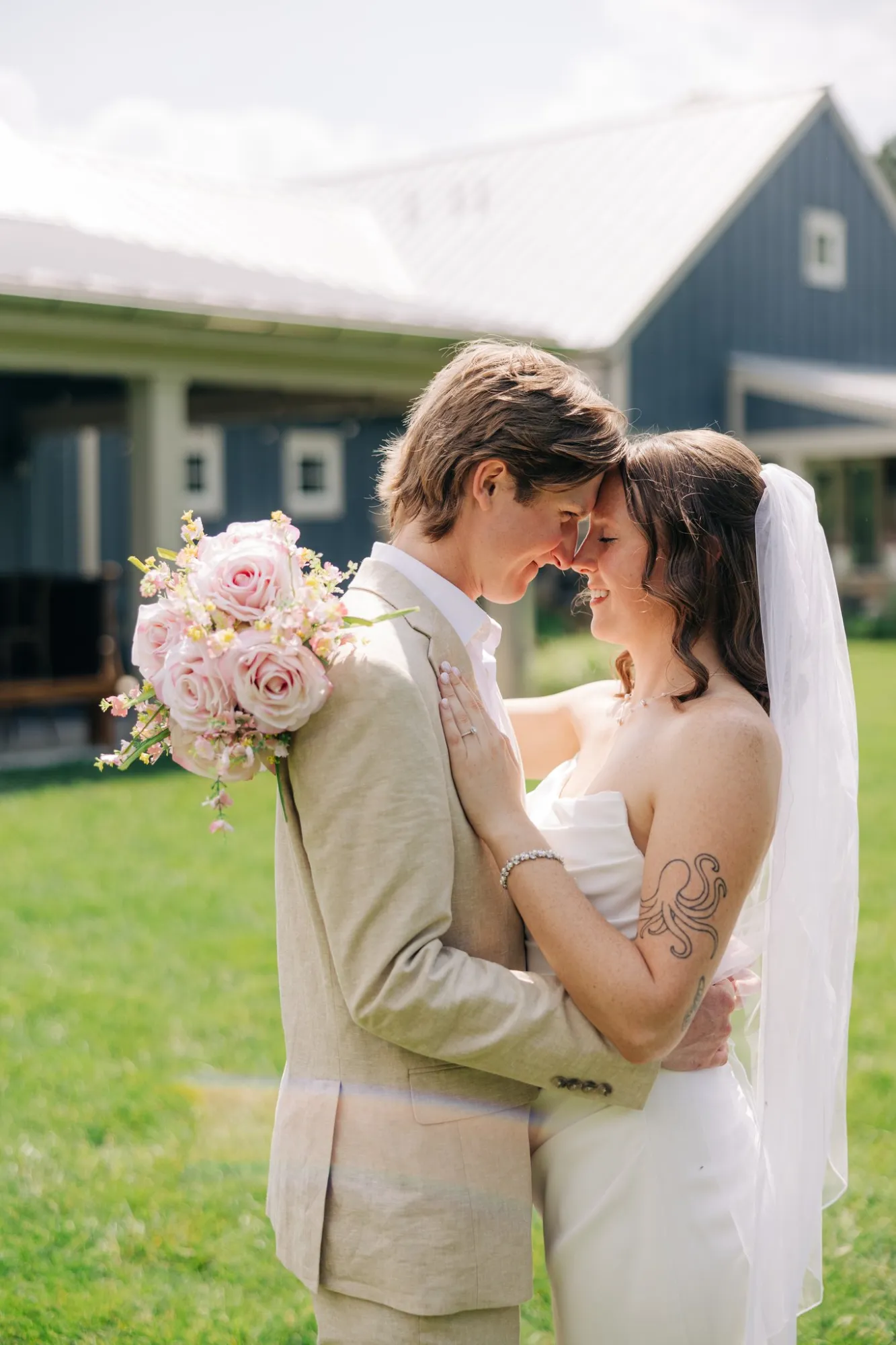 Bride and groom embracing outdoors with their foreheads touching, the groom holding a bouquet of pink roses, and the bride wearing a white dress and veil with an octopus tattoo on her upper arm.