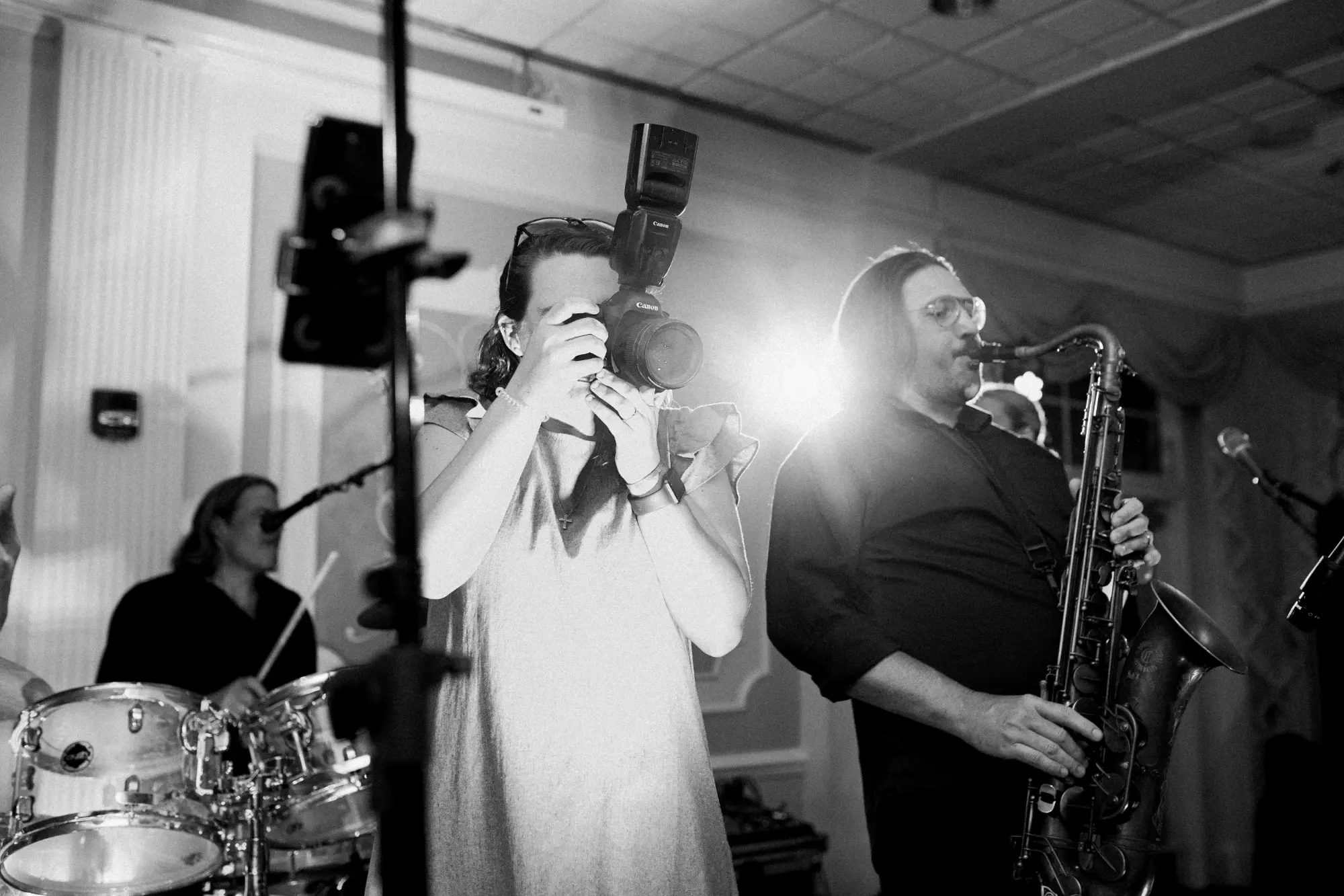 Black and white photo of a female photographer using a Canon camera with flash, standing next to a male saxophonist playing his saxophone, with a drummer and microphones in the background.
