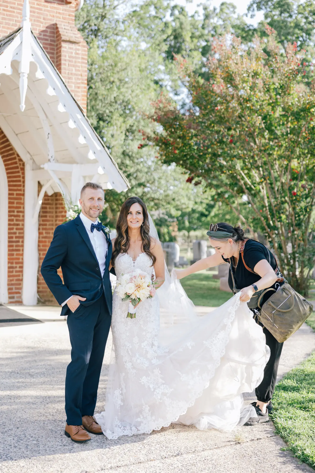 A bride and groom standing outside a building with red brick and white trim; the bride is holding a bouquet and wearing a lace wedding dress with a veil while a woman in black adjusts the train of her dress.