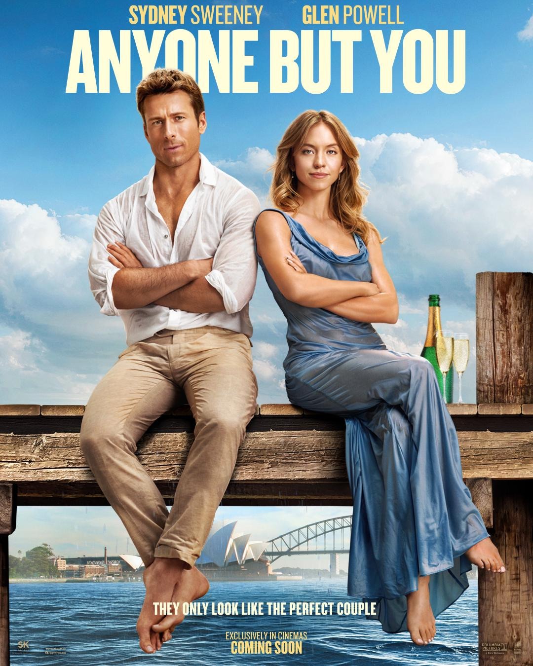 Movie poster for Anyone But You showing a man and woman sitting on a wooden dock with arms crossed, the Sydney Opera House and Harbour Bridge in the background under a blue sky, with two glasses and a champagne bottle beside the woman.