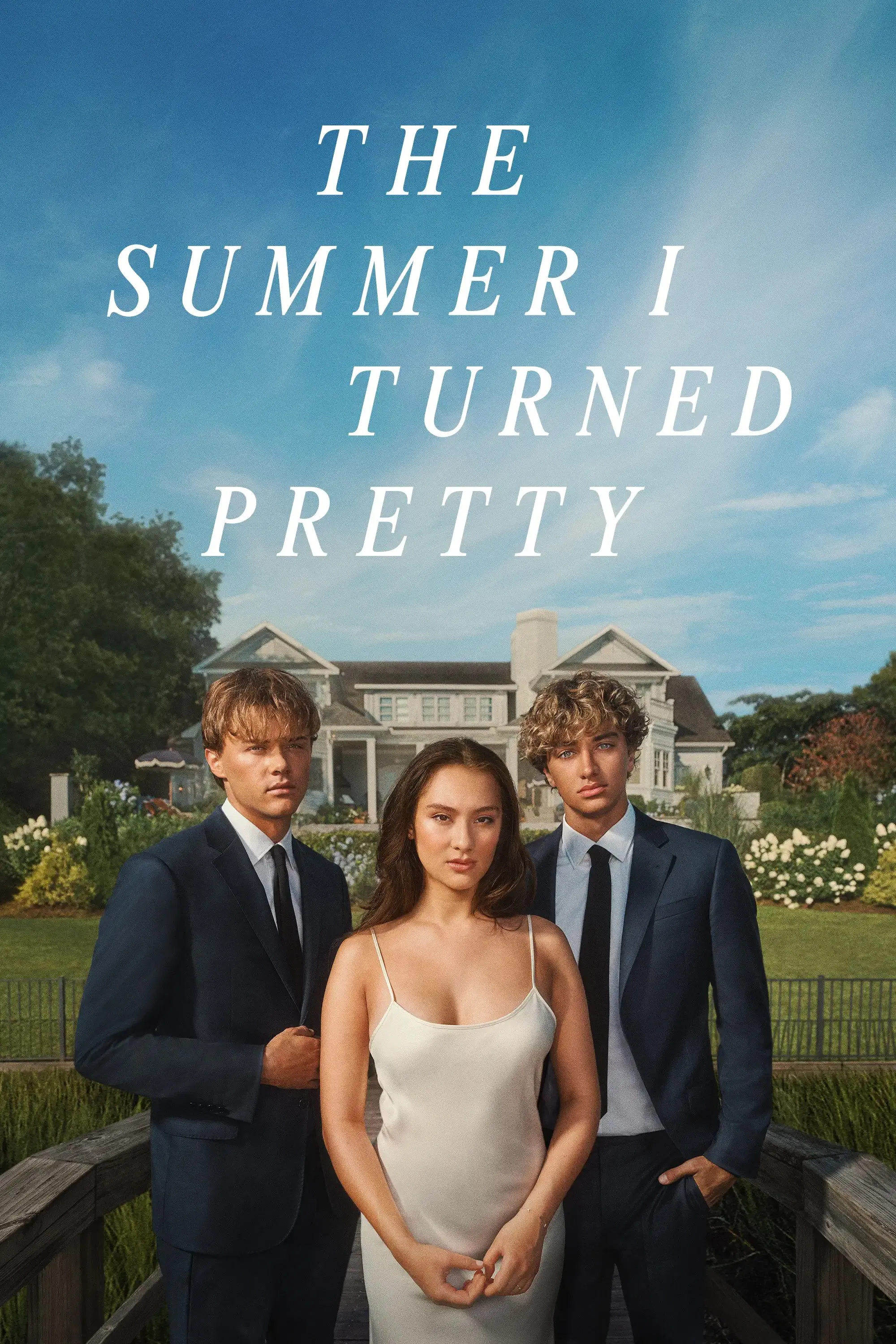 A young woman in a white dress stands between two young men in dark suits and ties on a wooden walkway, with a large house and garden in the background under a blue sky.