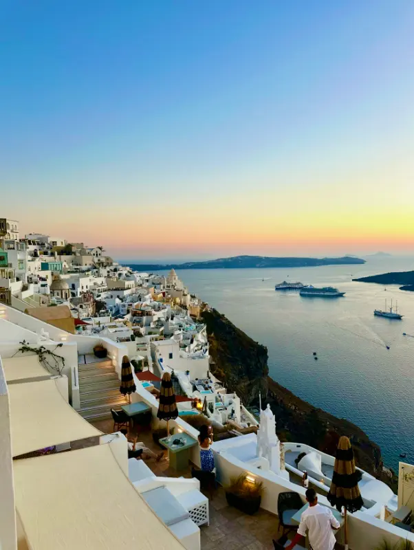 View of white buildings and terraces cascading down a cliffside at sunset in Santorini, Greece, overlooking calm blue sea with boats and distant islands under a clear sky.