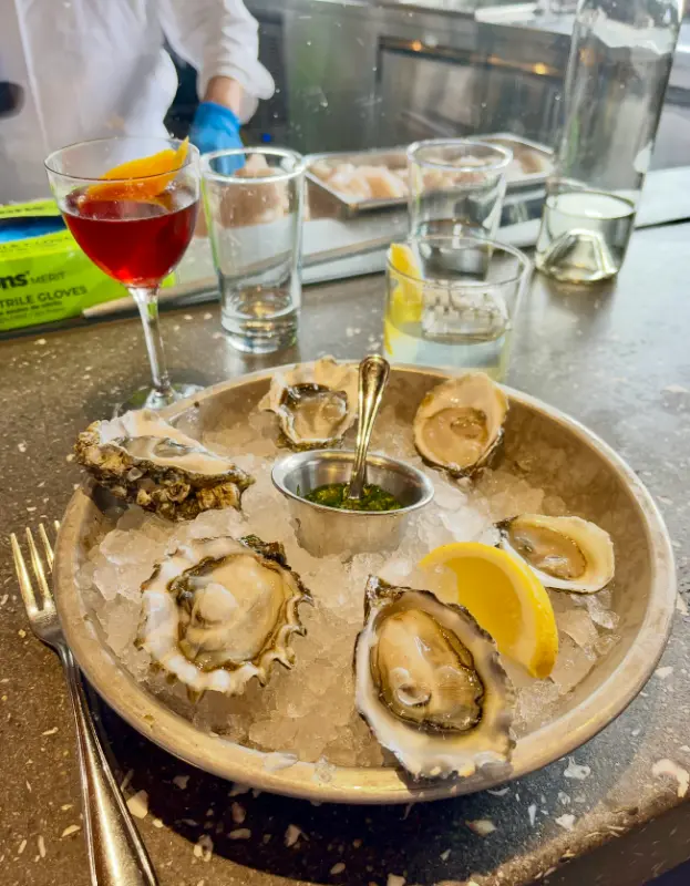 A plate of six raw oysters served on crushed ice with a lemon wedge and a small metal container of green sauce, placed on a gray countertop with a fork beside it. In the background are empty glasses, a cocktail with an orange peel garnish, and a person wearing a white shirt and blue gloves behind a glass partition.