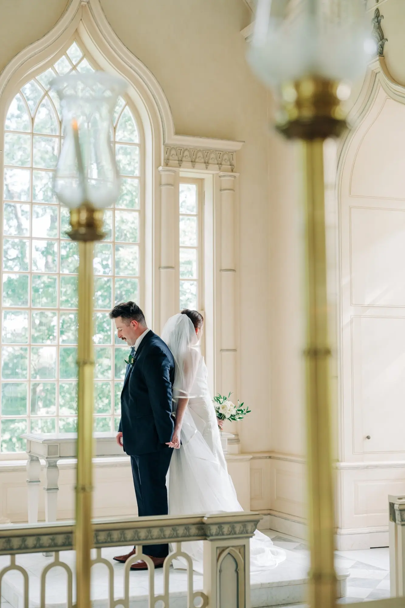 Bride in white wedding dress and veil holding a bouquet stands back-to-back, holding hands with the groom in a dark suit inside a sunlit room with large arched windows and ornate architectural details.