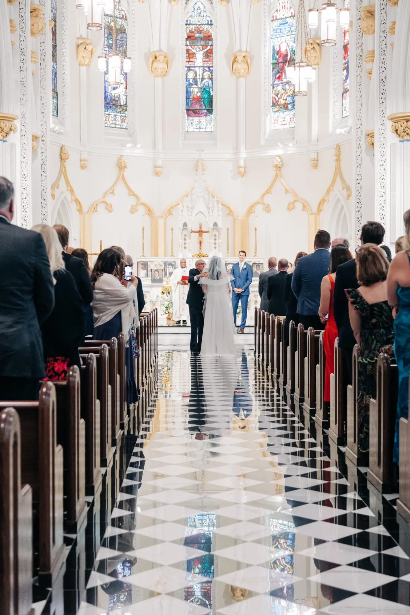 Bride walking down a black and white checkered aisle with a man in a dark suit in a bright church with stained glass windows, toward a groom in a blue suit standing at the altar with a clergyman, while guests watch from wooden pews.