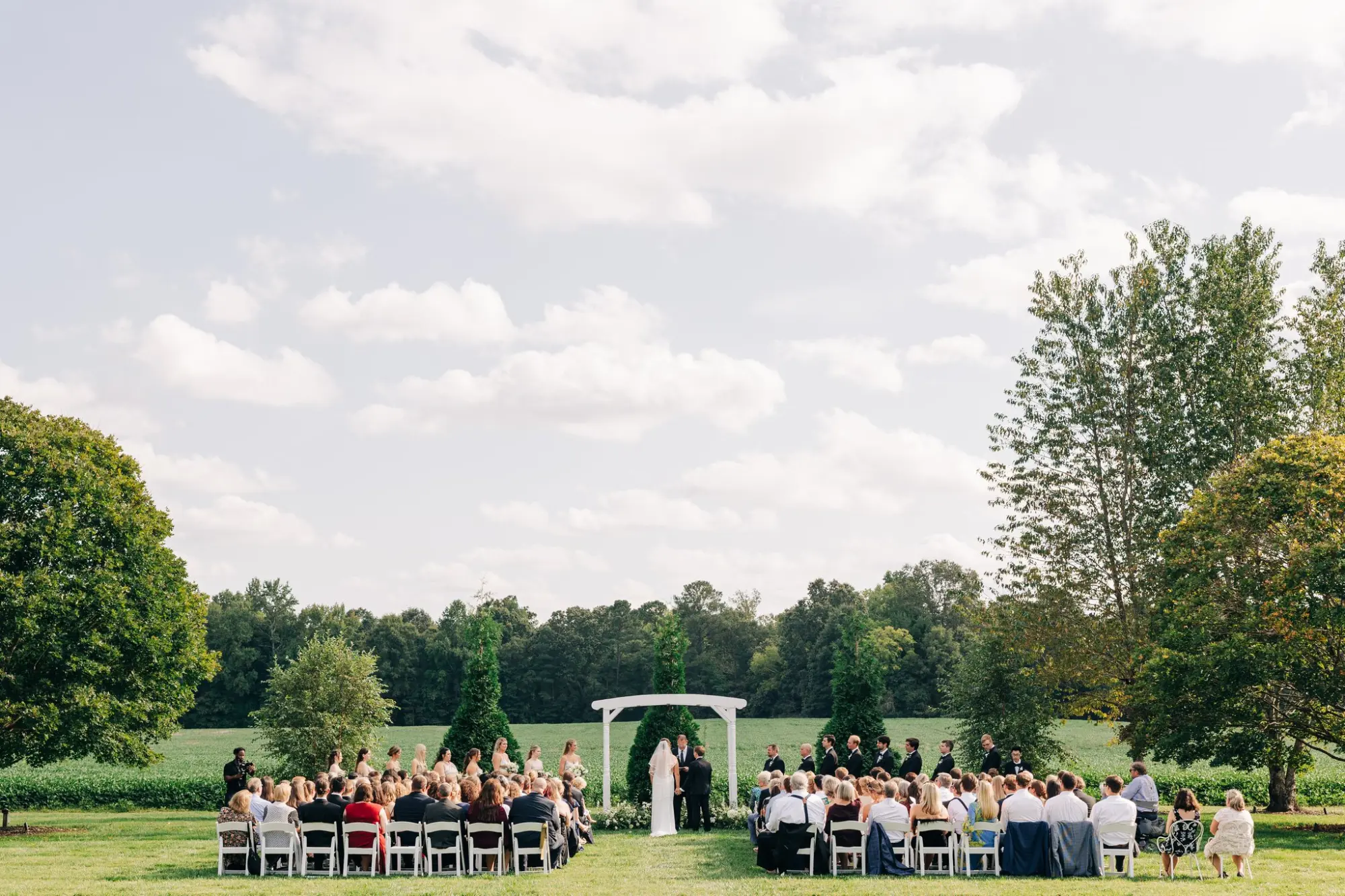 Outdoor wedding ceremony with bride and groom standing under a white arch, surrounded by bridesmaids in light dresses and groomsmen in black suits, guests seated on white chairs on both sides, green trees and fields in the background under a partly cloudy sky.