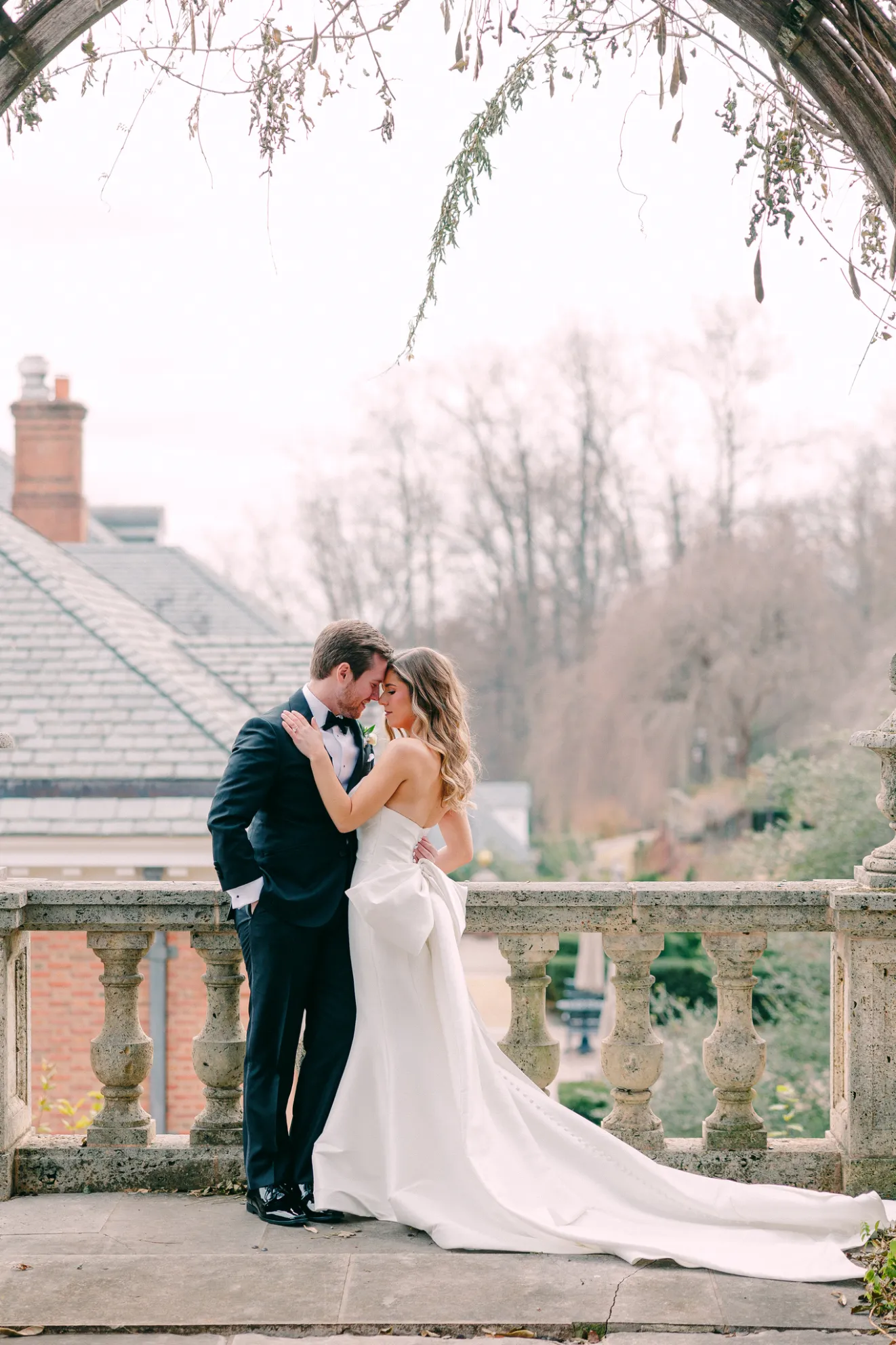 Bride in a strapless white gown with a long train and groom in a black tuxedo standing close and embracing under a rustic wooden arch with hanging greenery on a stone balcony overlooking a blurred outdoor garden and trees.