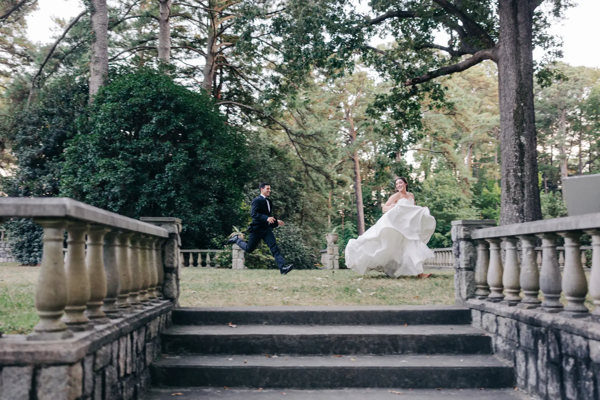 Bride in white dress and groom in blue suit holding hands and walking barefoot through a vineyard with leafless grapevines and autumn trees in the background.