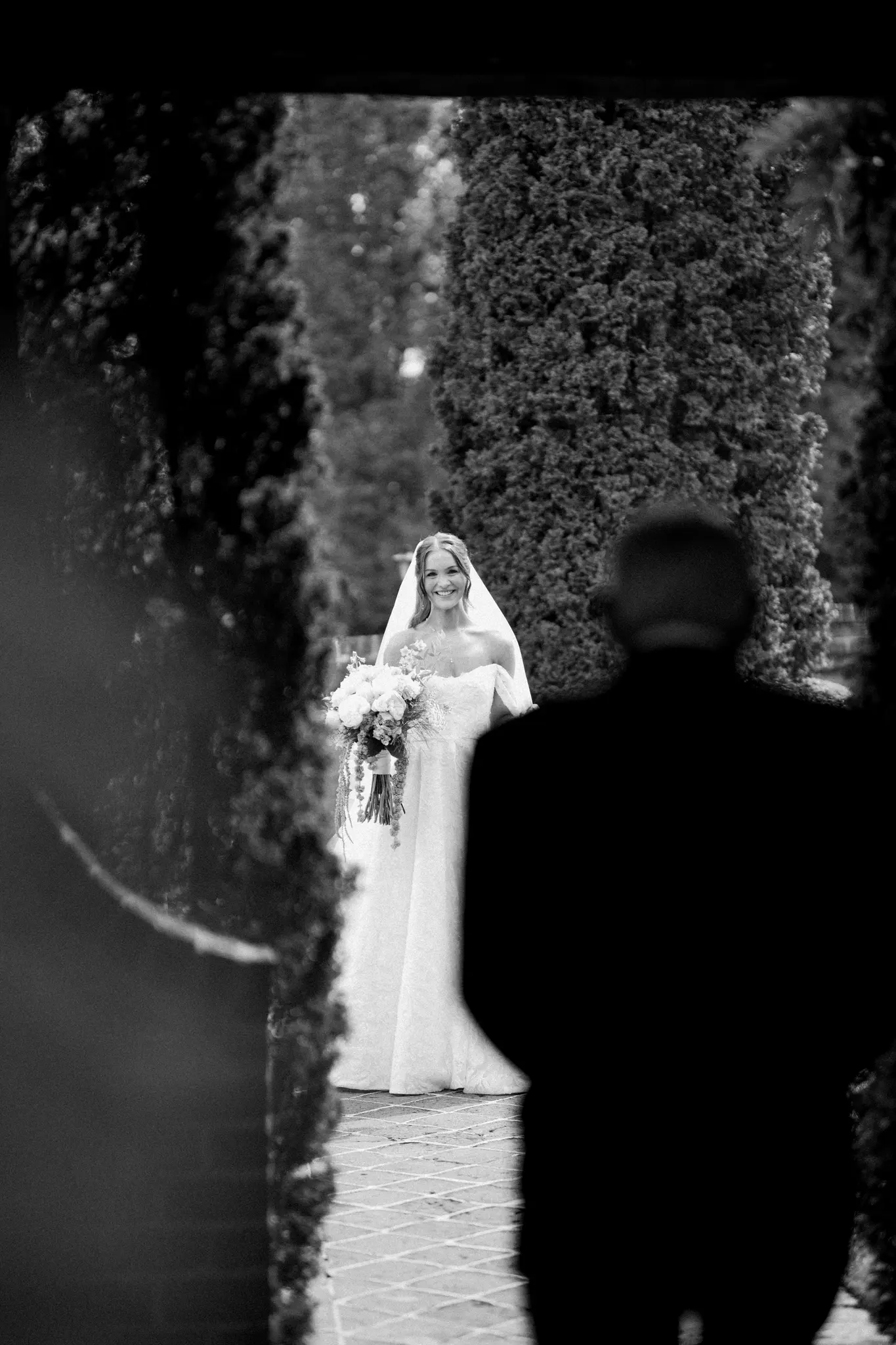 Black and white photo of a smiling bride in a strapless wedding dress and veil, holding a bouquet, walking outdoors toward a person in the foreground whose back is to the camera.