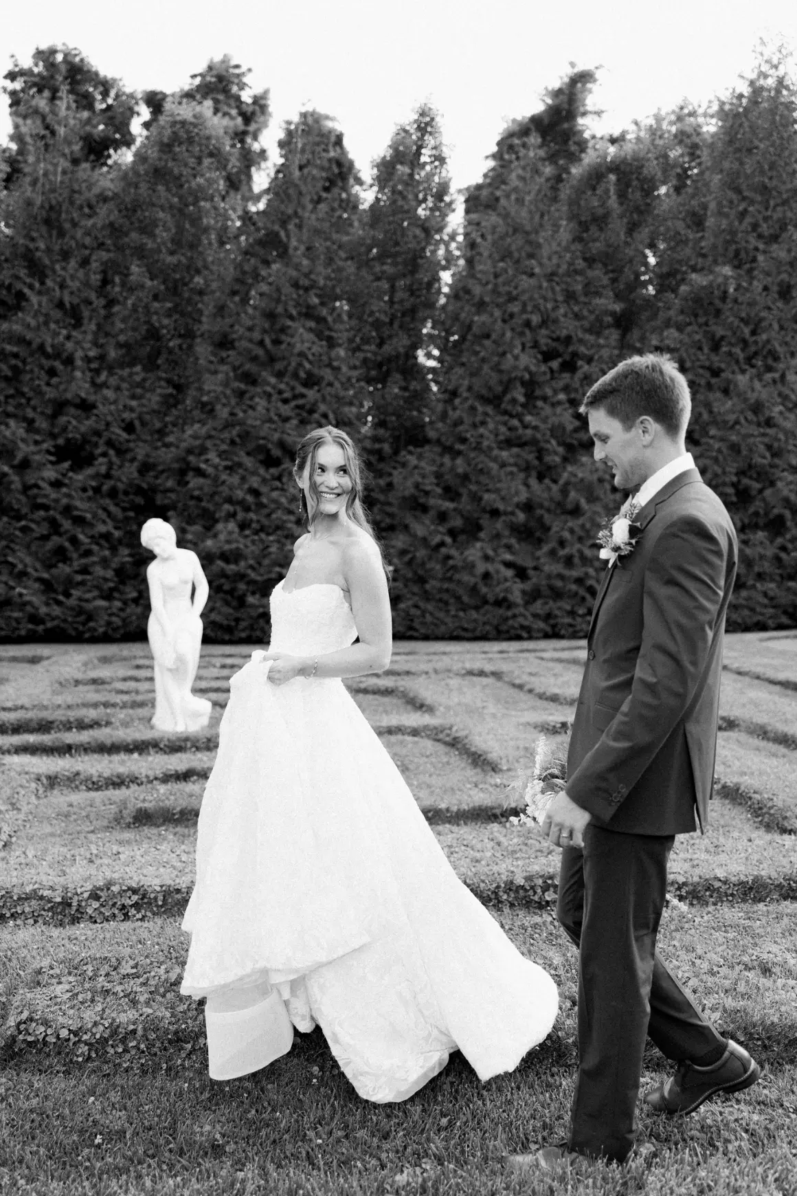 Black and white photo of a bride in a strapless wedding gown smiling and holding her dress, standing on grass with a groom in a suit looking at her, in a garden with a statue and tall hedges in the background.