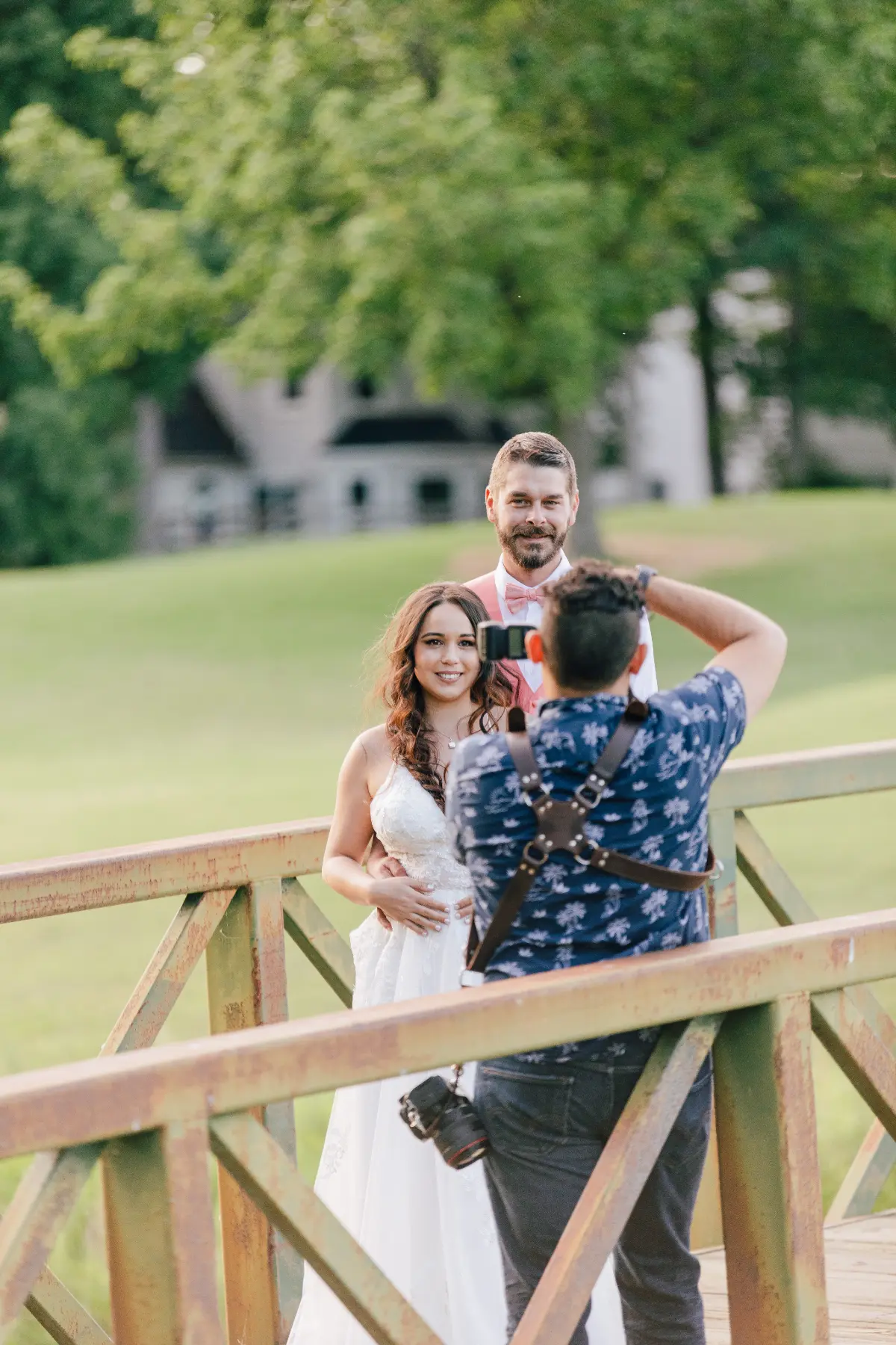Photographer taking a picture of a smiling bride in a white dress and a groom in a pink vest and bow tie on a rustic wooden bridge with green grass and trees in the background.