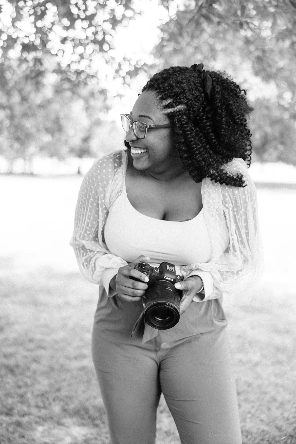 Smiling woman with curly hair and glasses stands outdoors holding a Canon camera, looking to her left.