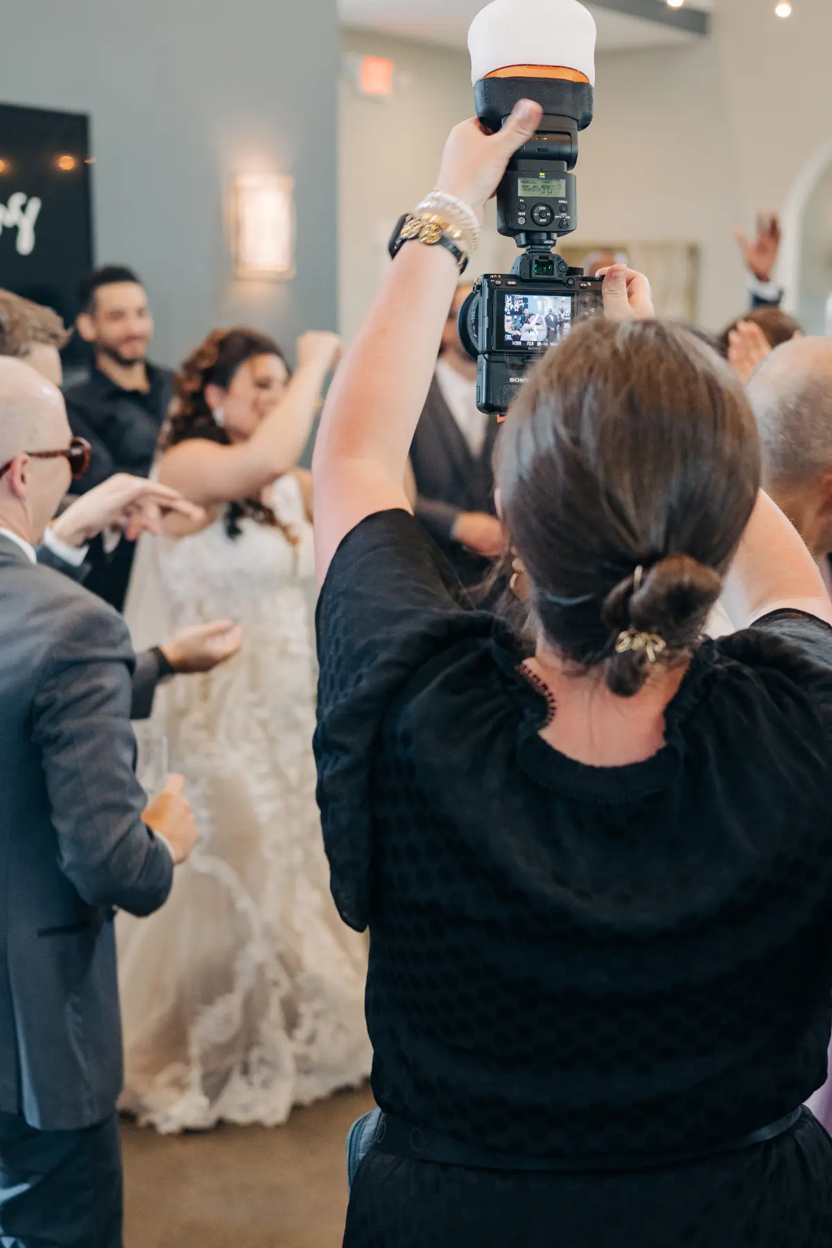 Photographer with hair in a bun and wearing a black dress takes a photo with a camera and flash at a wedding, capturing a bride in a white lace gown and guests celebrating.