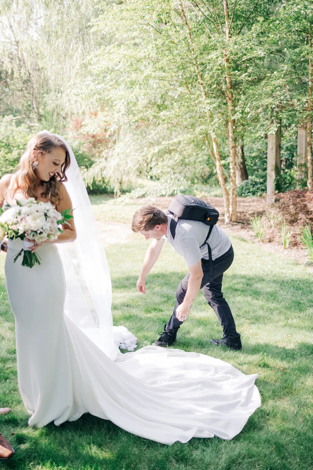 Bride in a white wedding dress holding a bouquet smiles as a man with a backpack adjusts the train of her dress on green grass outdoors with trees in the background.