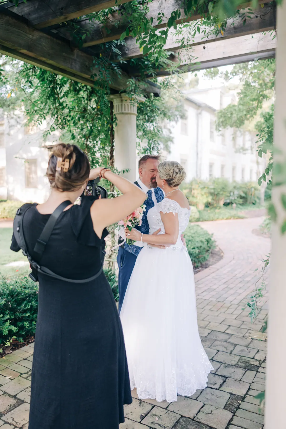 Photographer in a black dress taking a picture of a bride and groom standing under a wooden pergola wrapped in greenery, with a paved path and white building in the background.