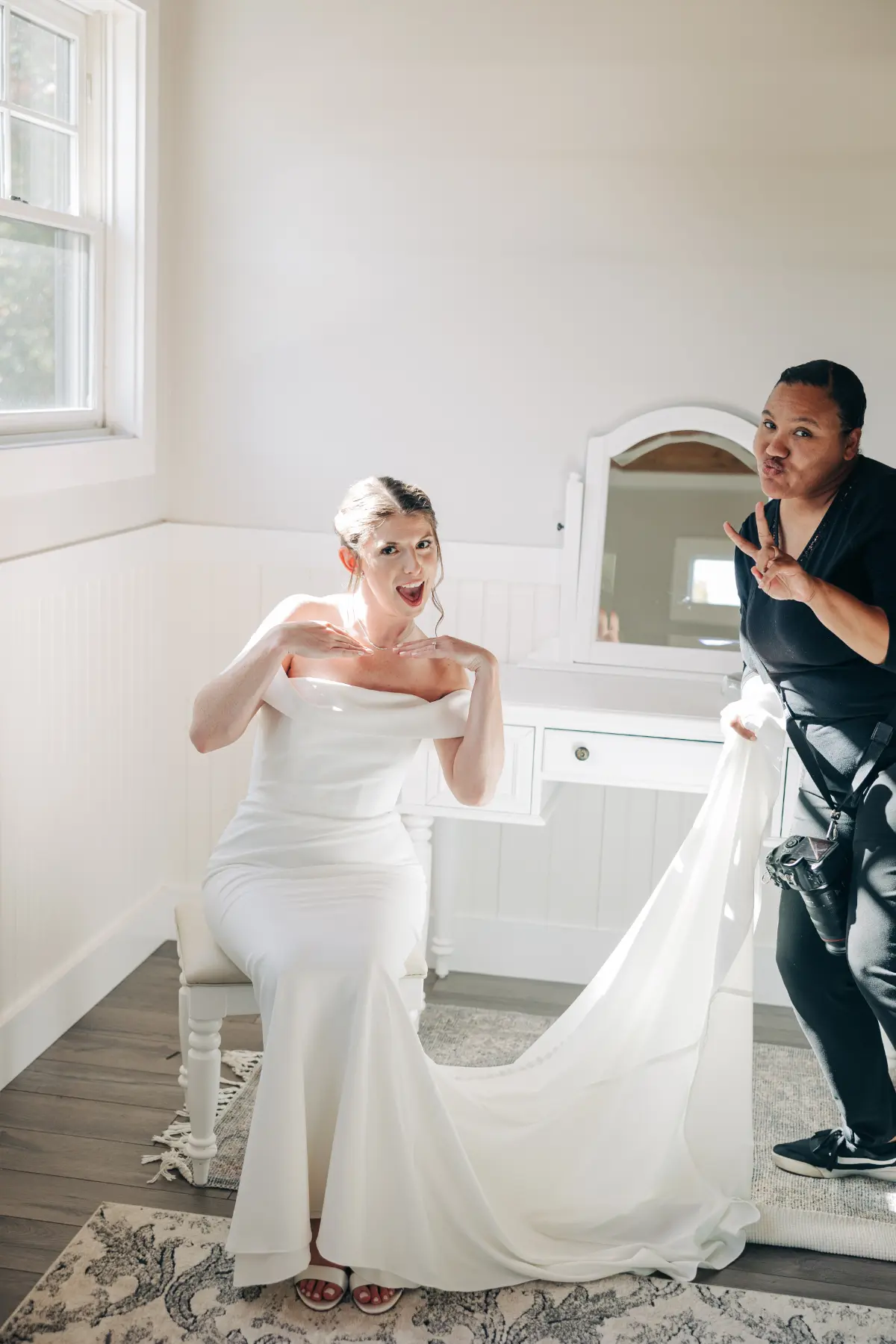 A bride in an off-the-shoulder white wedding dress sits on a bench smiling and posing playfully with her hands near her chin, while a woman stands beside her holding the train of the dress and making a peace sign with her fingers.