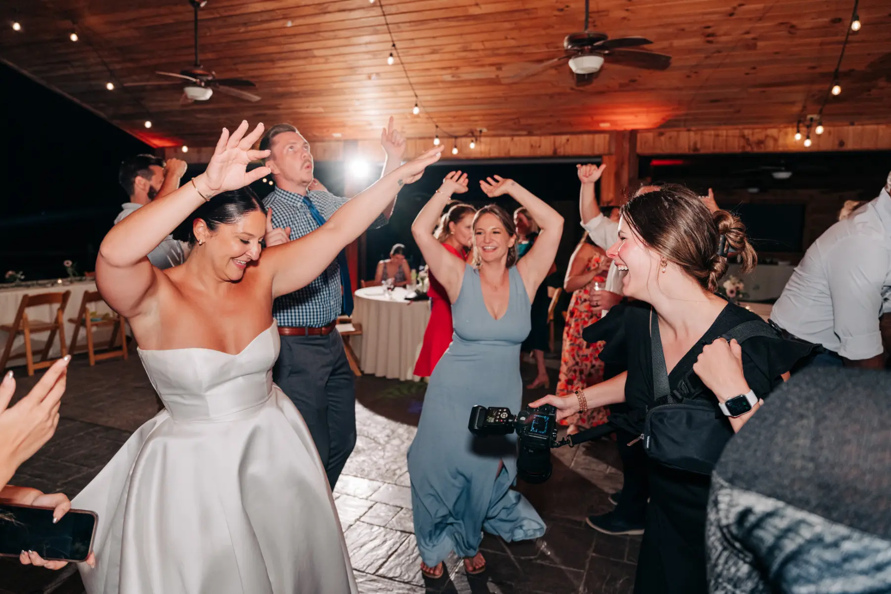 People dancing and smiling at a wedding reception, including a bride in a strapless white dress, a woman in a blue dress, and a photographer holding a camera, under a wooden ceiling with string lights.