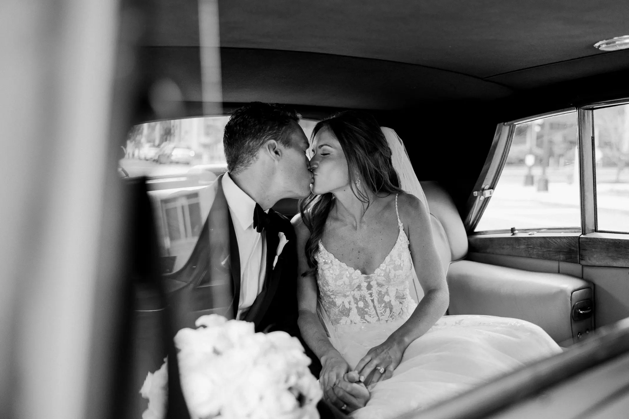 Black and white photo of a bride and groom kissing inside a car, holding hands, with the bride wearing a lace wedding dress and veil.