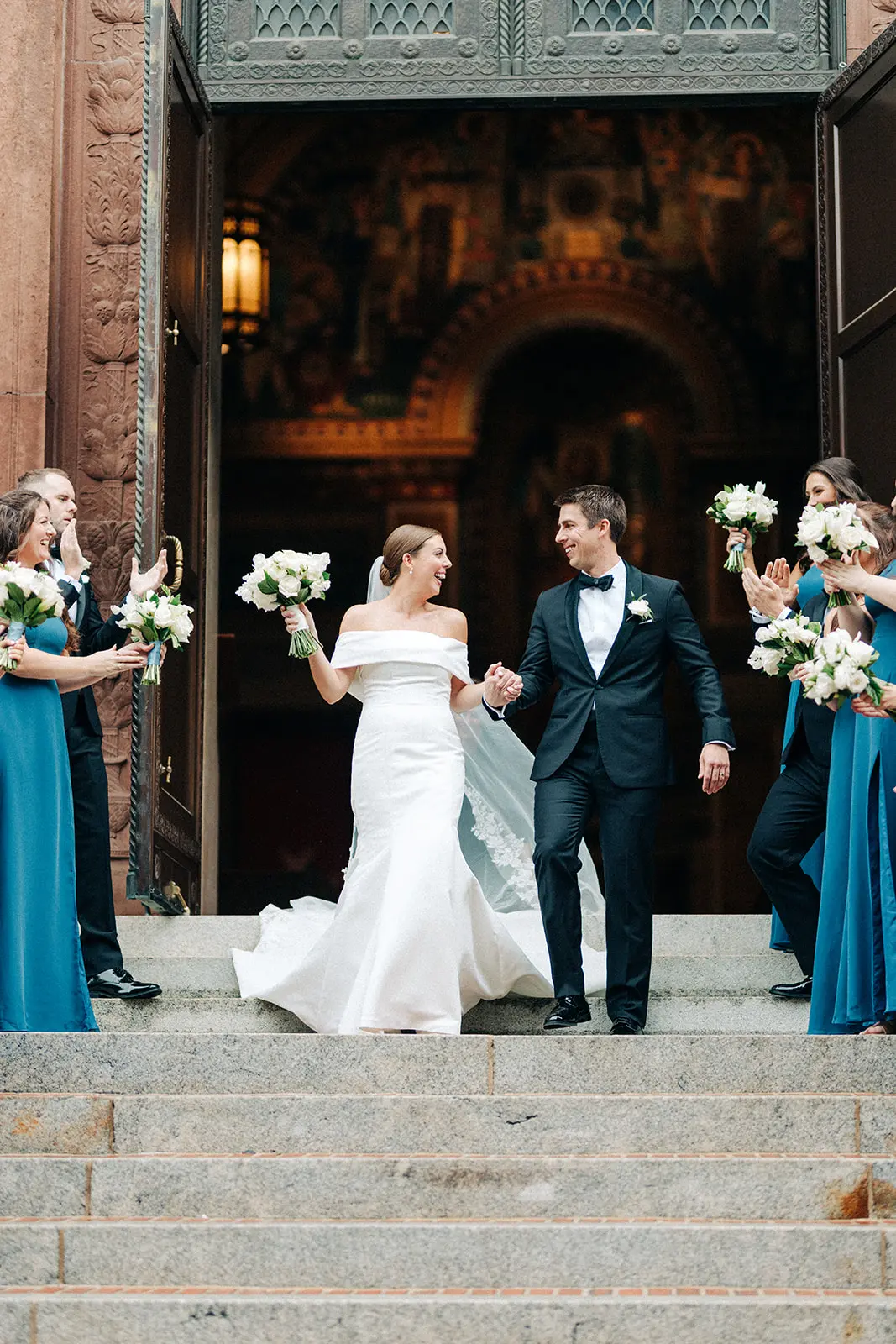 Bride in white off-shoulder gown holding a bouquet and groom in black tuxedo holding hands and smiling as they walk down stone steps outside a large open doorway, with bridesmaids and groomsmen in teal dresses and black suits clapping and holding bouquets on either side.