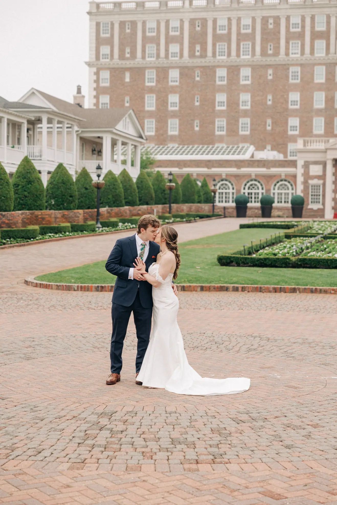 Bride and groom dressed in wedding attire sharing a kiss outdoors on a patterned brick courtyard with manicured gardens and a large multi-story brick building in the background.