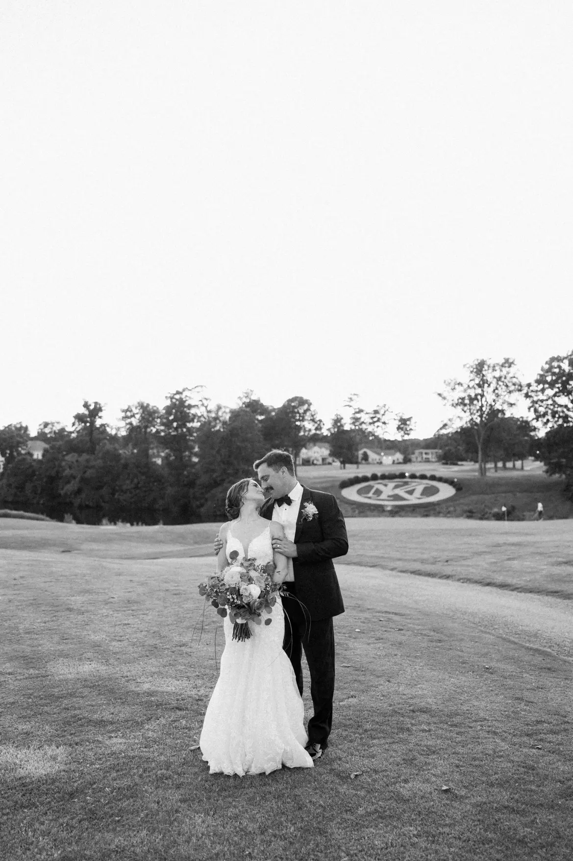 Bride and groom standing closely outdoors with the bride holding a bouquet of pink roses and other small flowers, the bride wears a white dress and veil, and the groom wears a light beige suit with a green pocket square.