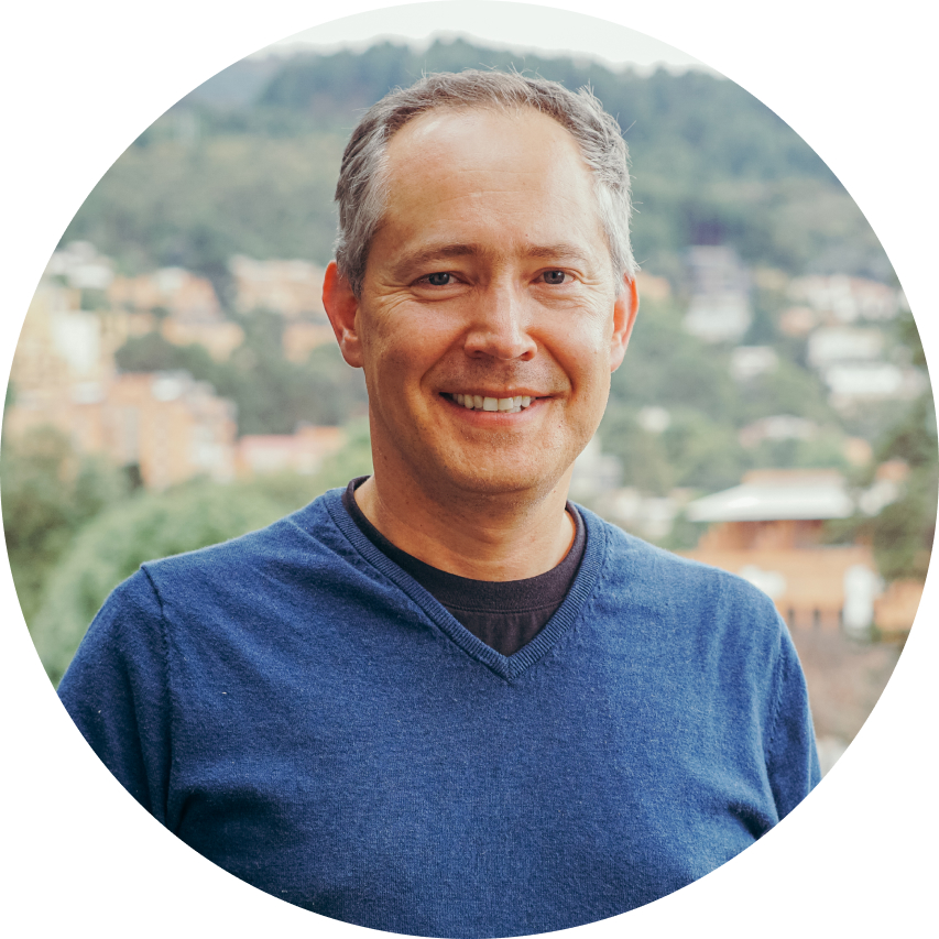 Smiling middle-aged man in blue sweater with blurred town and forest background.