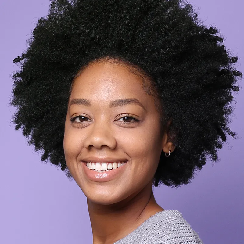 Smiling woman with natural curly hair wearing a gray sweater against a purple background.