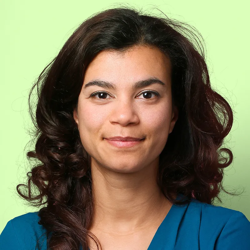Smiling woman with curly dark hair wearing a blue top against a light green background.