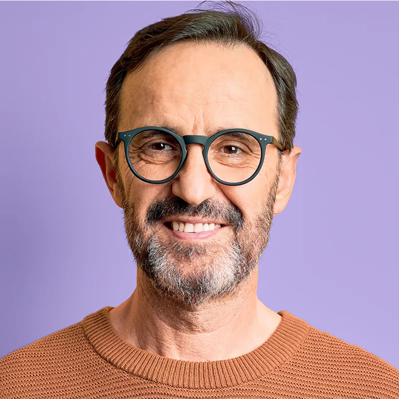 Smiling middle-aged man with glasses and a beard wearing a brown sweater against a purple background.