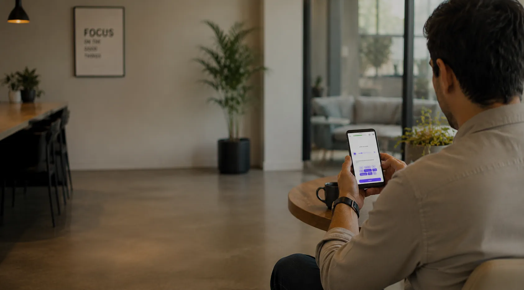 Man sitting at a round table holding a smartphone displaying an app interface in a modern room.