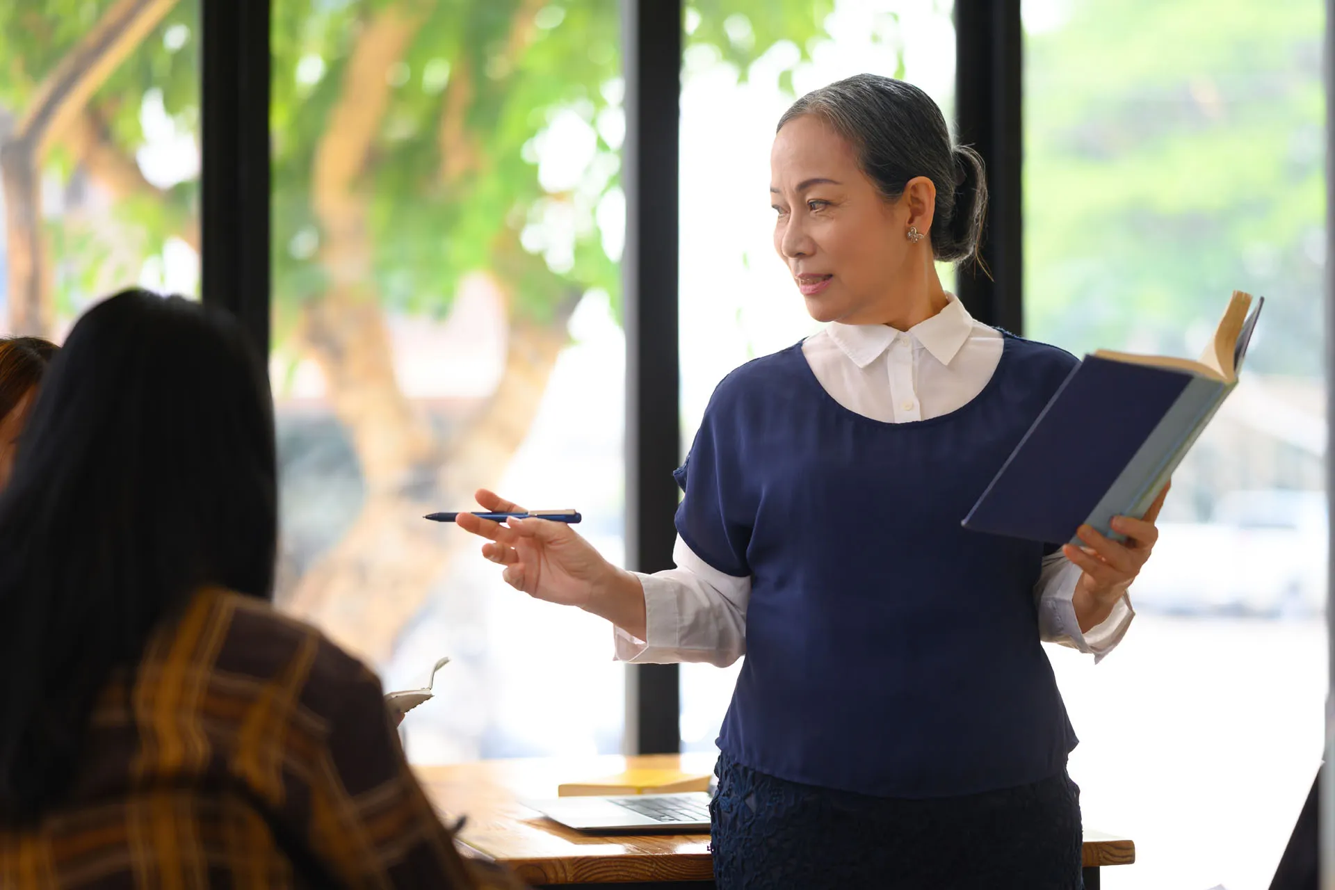 Mature female lecturer holding a book and pen, speaking to students in a classroom with large windows.