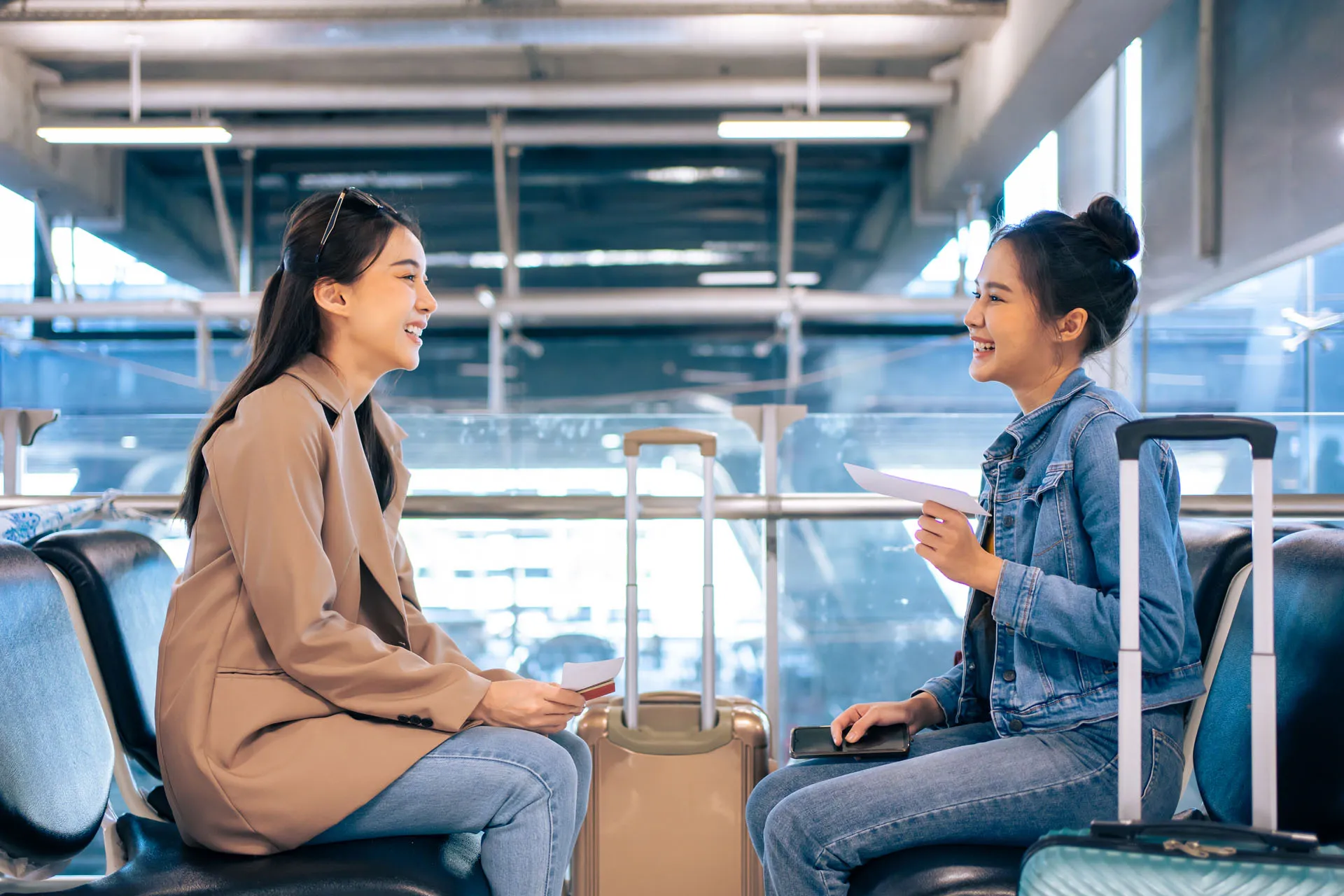 Two young women with luggage smiling and holding boarding passes while sitting in an airport waiting area.