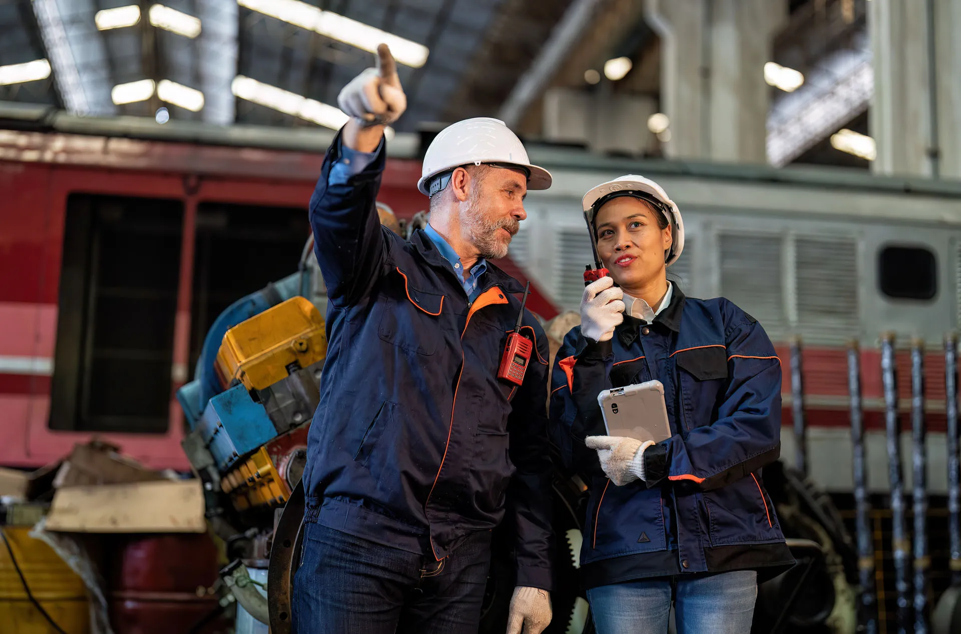 Two engineers wearing hard hats and jackets communicate with walkie-talkies in an industrial setting.