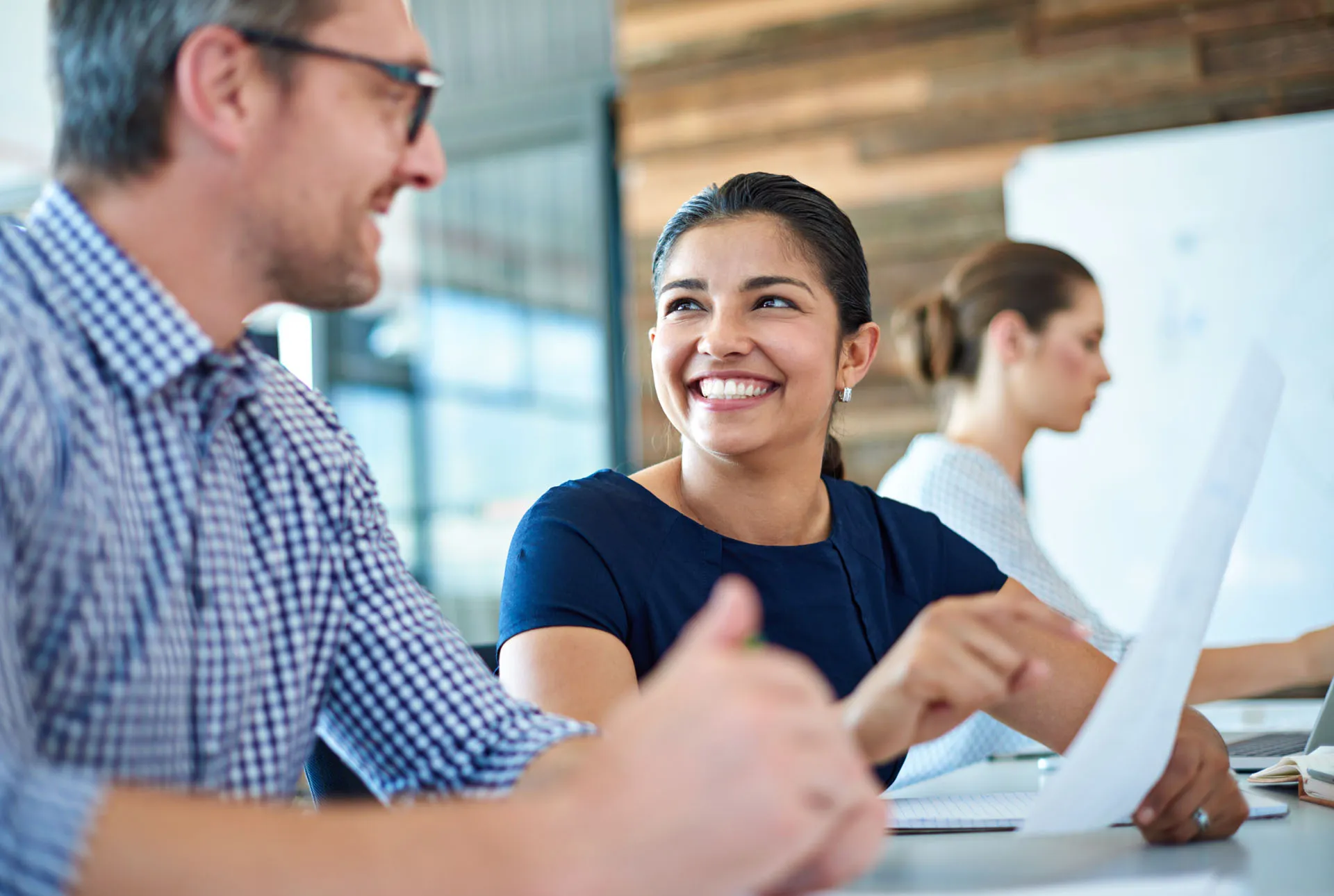 Smiling woman holding paper and talking to a man in a casual office setting.