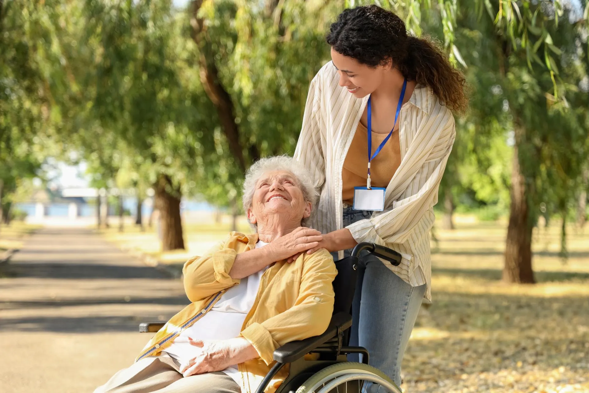 Young caregiver smiling and pushing elderly woman in wheelchair on a tree-lined path outdoors.