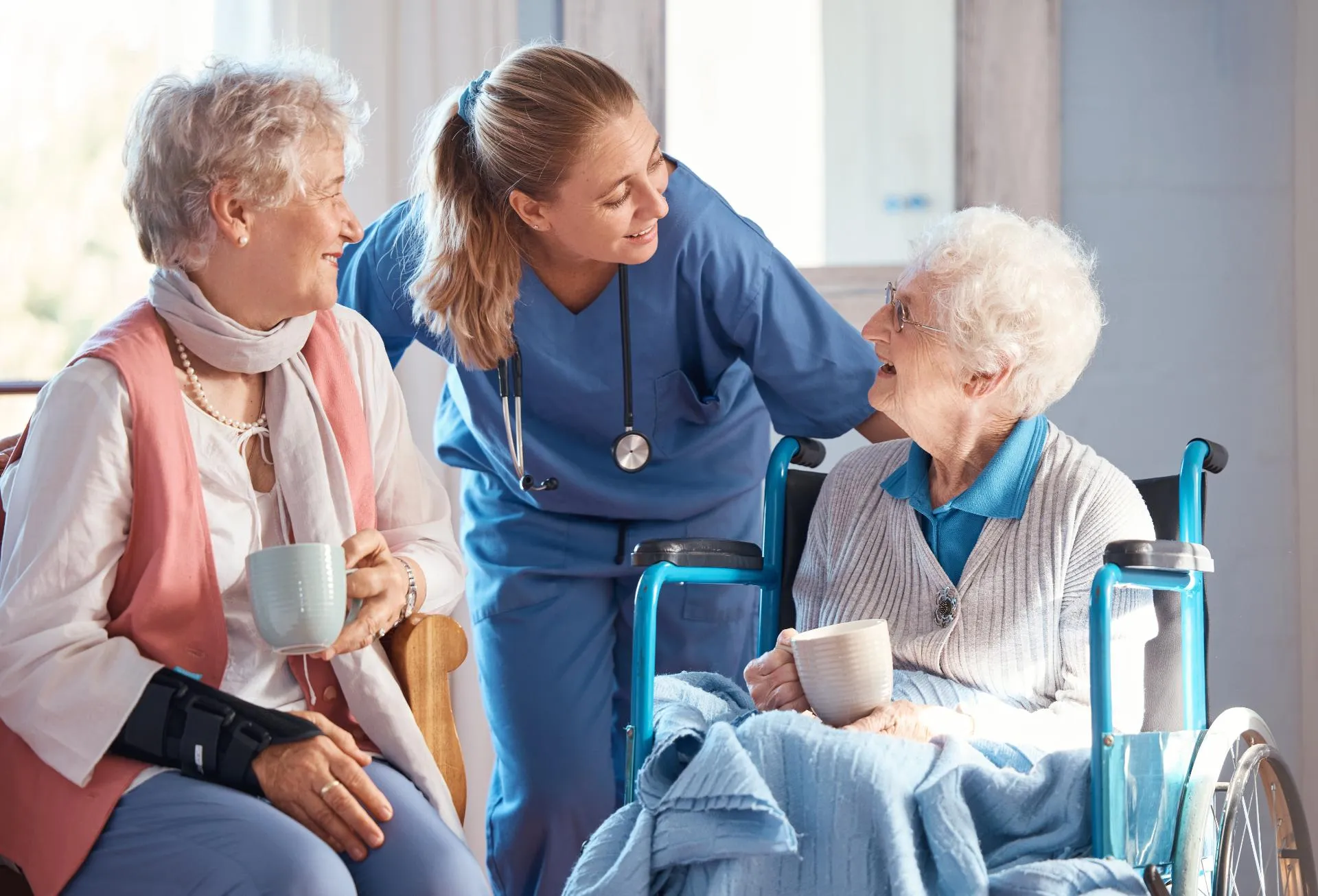 Nurse in blue scrubs talking with two elderly women, one seated in a wheelchair holding mugs.
