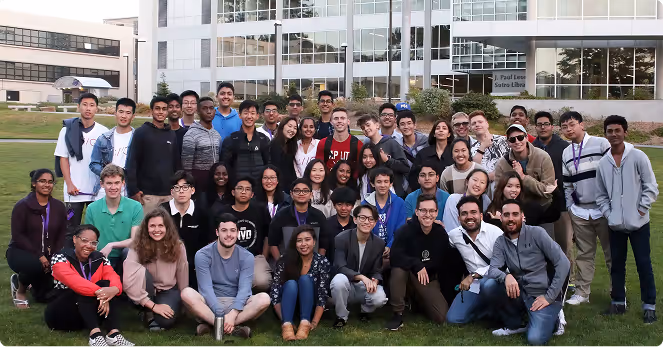 Large group of young adults posing together on grass outside a modern building.