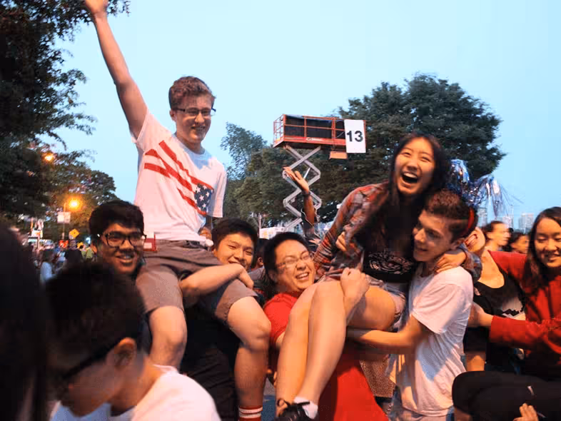 Group of young people outdoors, two being lifted up, one wearing USA flag shirt, all smiling and happy.