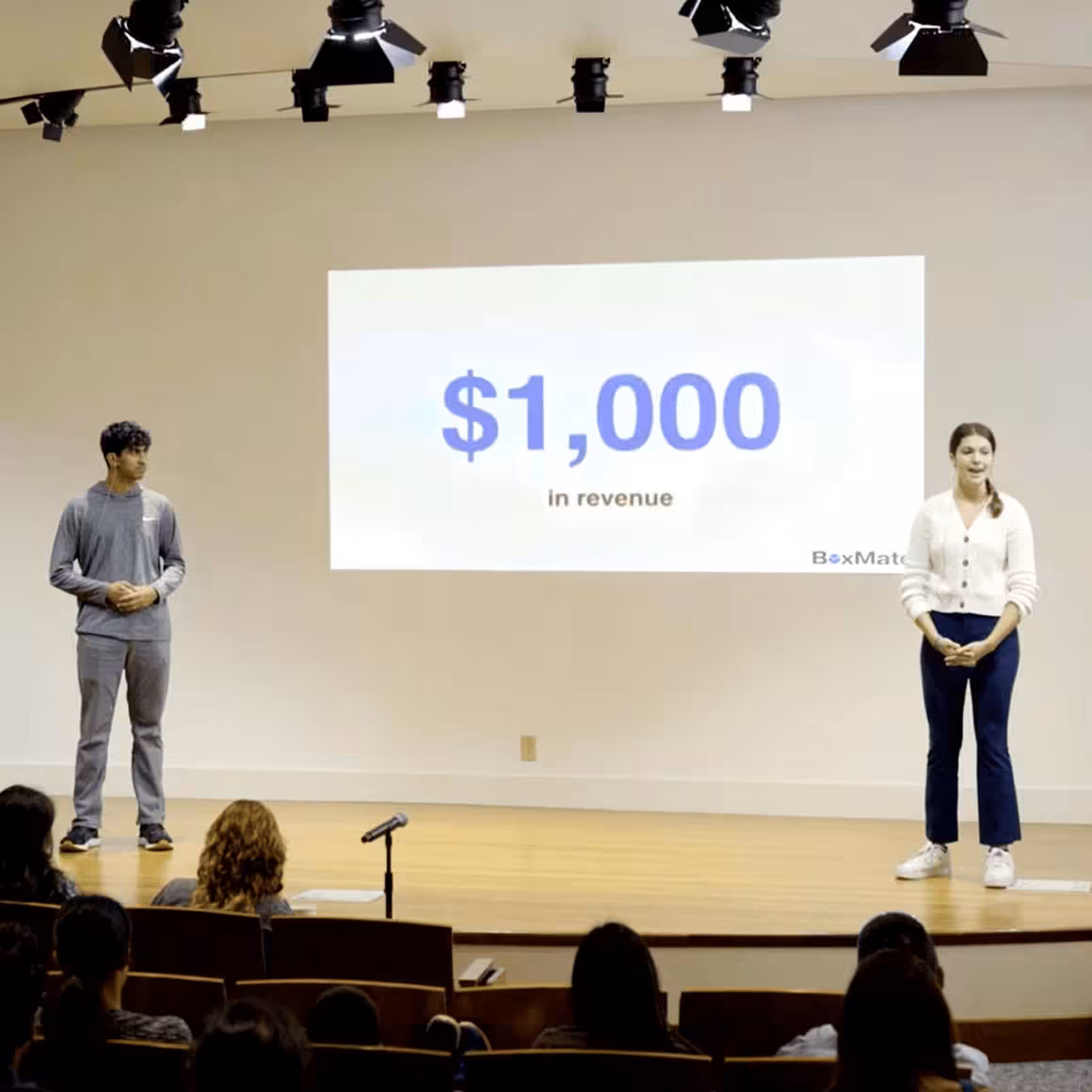 Two teens standing on stage presenting with a screen showing $1,000 in revenue behind them.