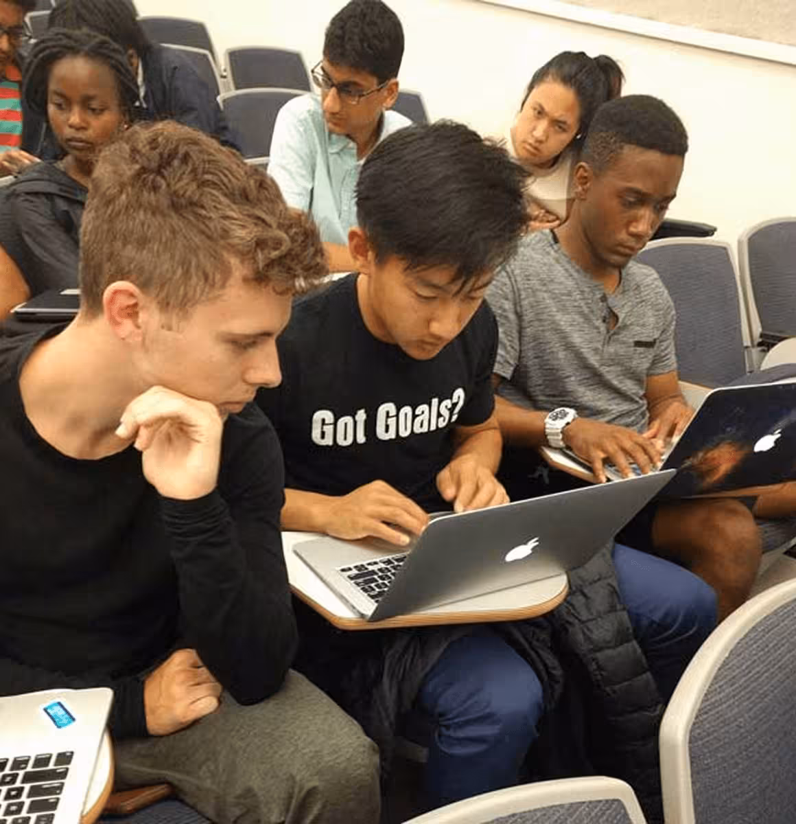Group of diverse students working on laptops in a classroom with desks and chairs.