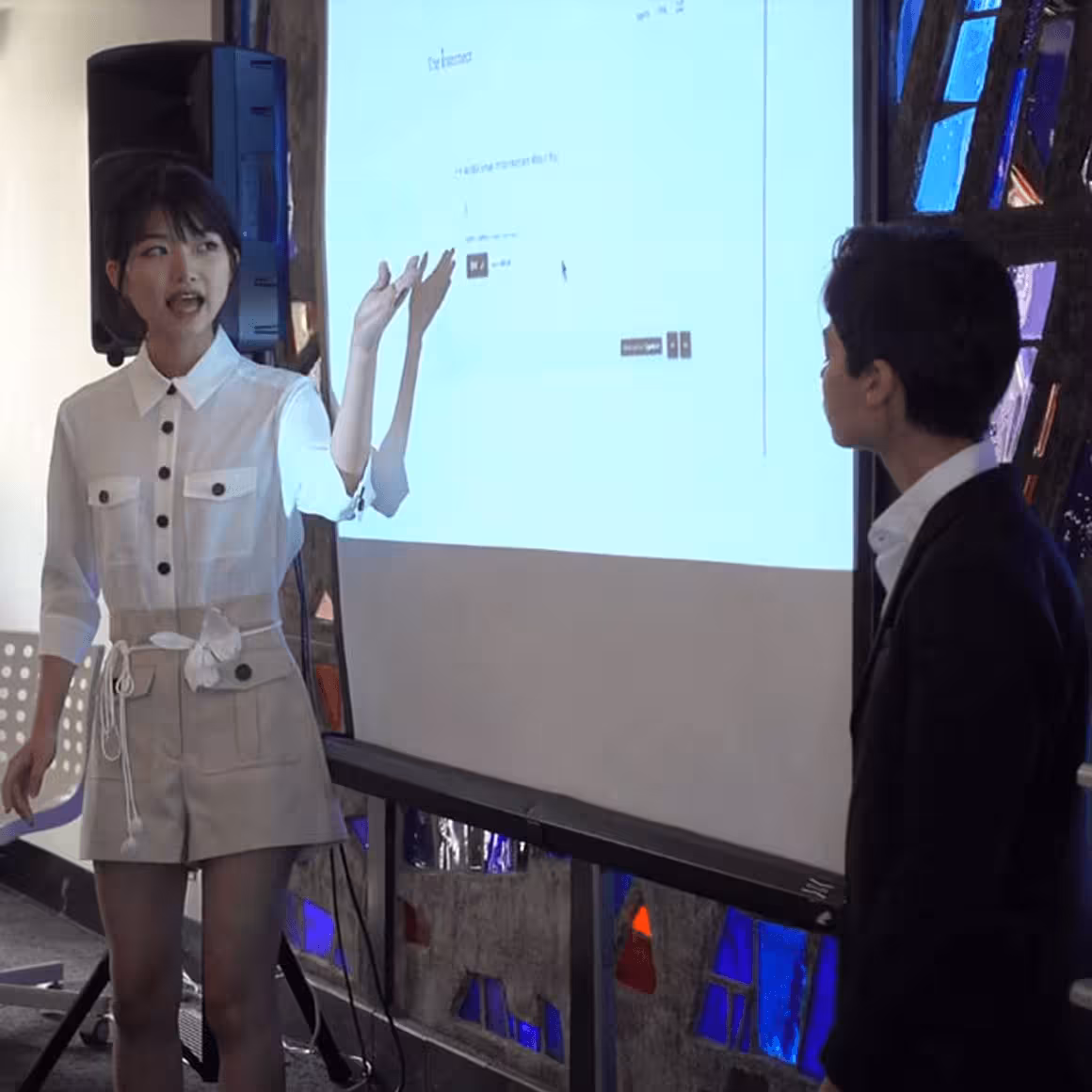 Woman in white shirt and shorts giving a presentation to a man in a suit.
