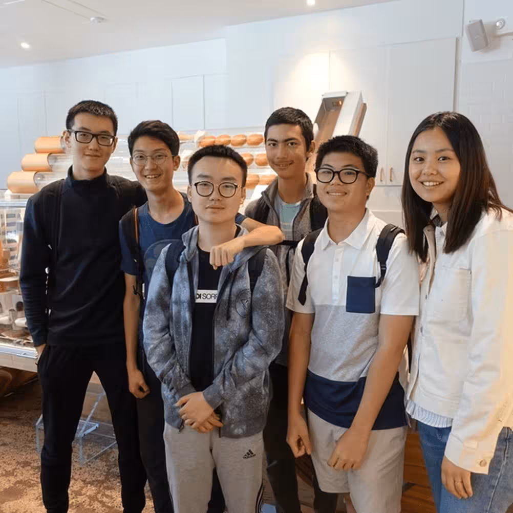 Group of six young students smiling in a bakery or café with bread visible in the background.