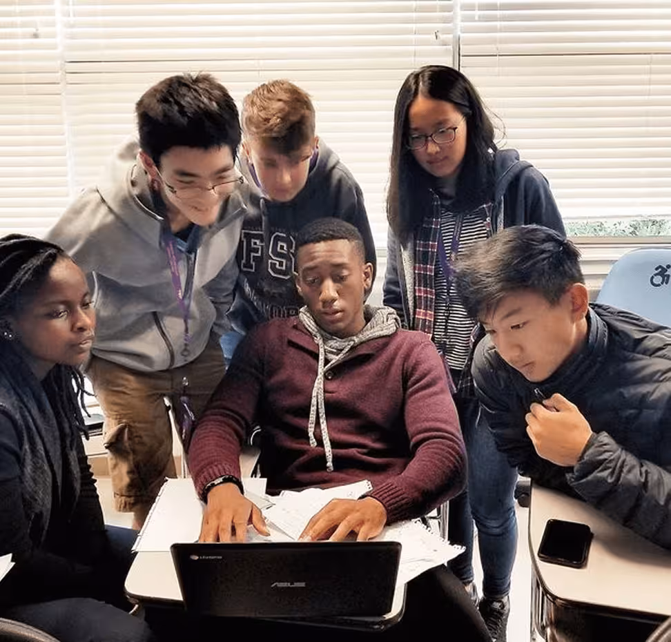 Six diverse students gathered around a laptop, engaged in group study in a classroom with blinds.